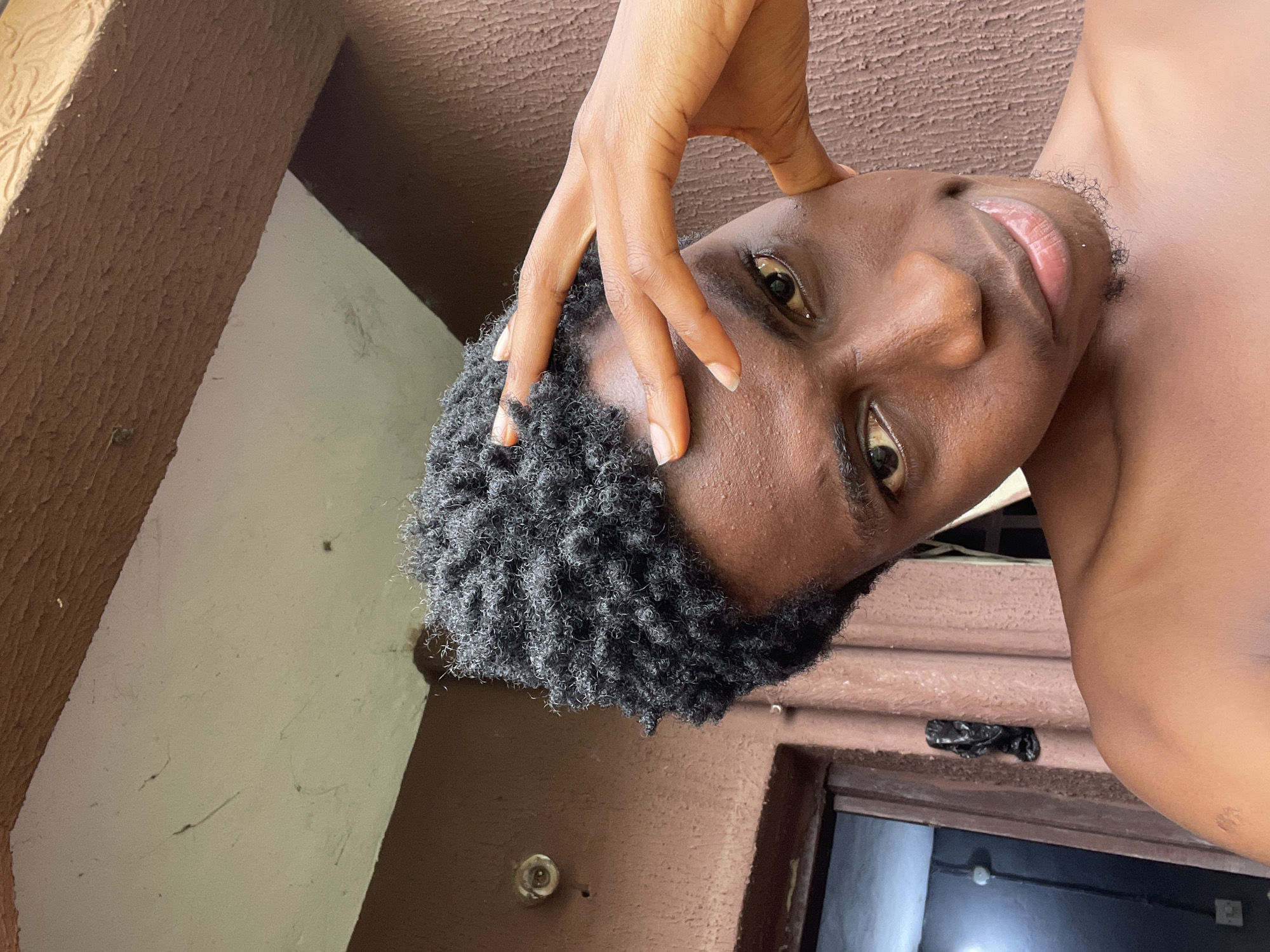 A close-up, waist-up, outdoor selfie shows a young Black male with dark, curly hair and brown skin. He is looking directly at the camera with a slight smile and is touching his forehead with his right hand. The background consists of textured brown walls and a lighter colored ceiling or overhang. A doorway with a visible interior is to the left of the subject. The scene is consistent with an outdoor residential area in Nkpor, Nigeria.