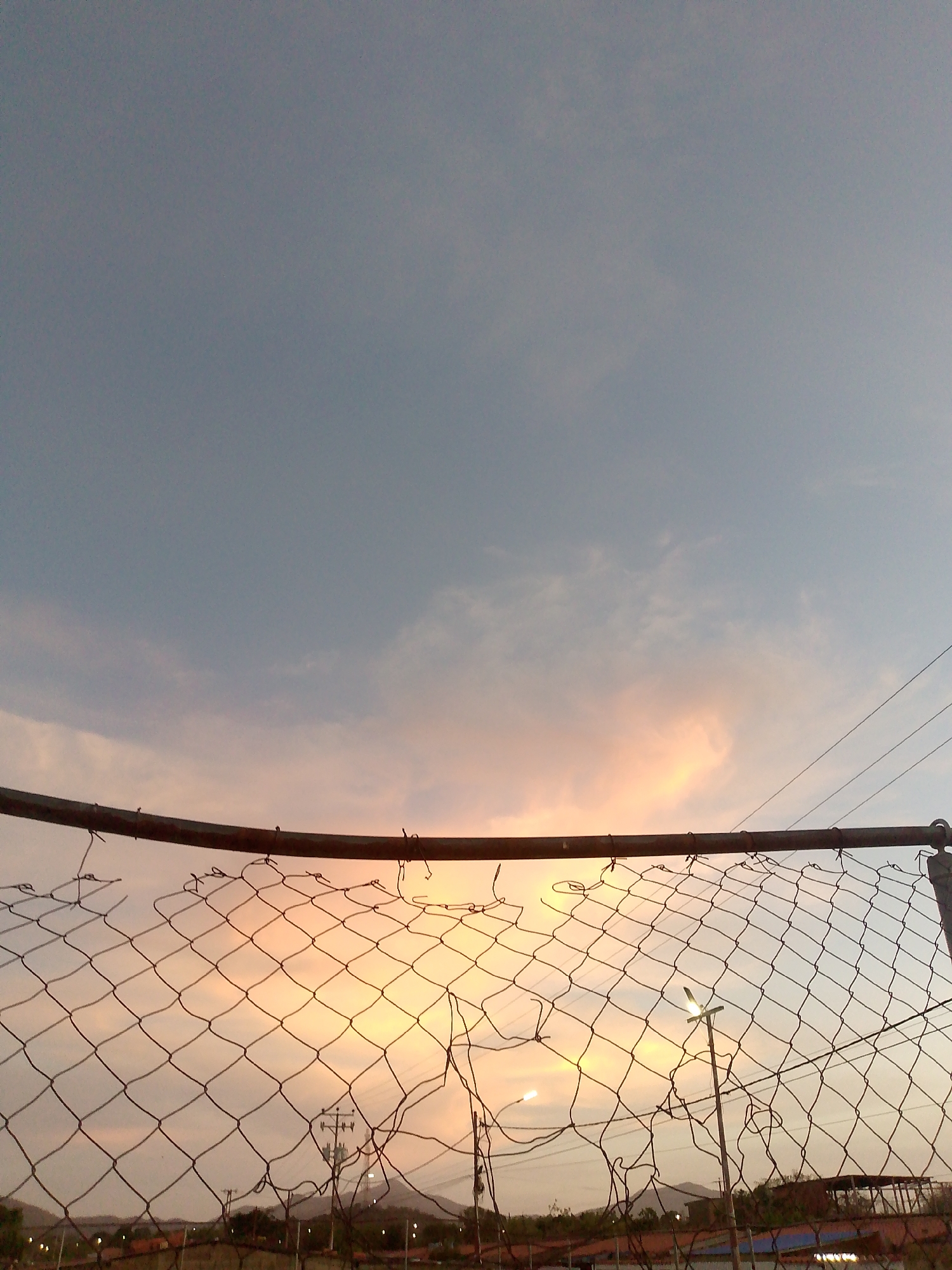 A low-angle, medium shot depicts a wire mesh fence in the foreground, with the sky and a cityscape visible through the fence. The sky is mostly blue, with some clouds tinged with orange and pink due to the sunset. Overhead power lines cross the upper right quadrant of the image. The wire fence is somewhat damaged with broken sections. In the middle ground, a few street lights and a building with a distinct metal frame are visible. In the background, a low mountain range forms the horizon. The city of Altagracia, Venezuela is the presumed location.
