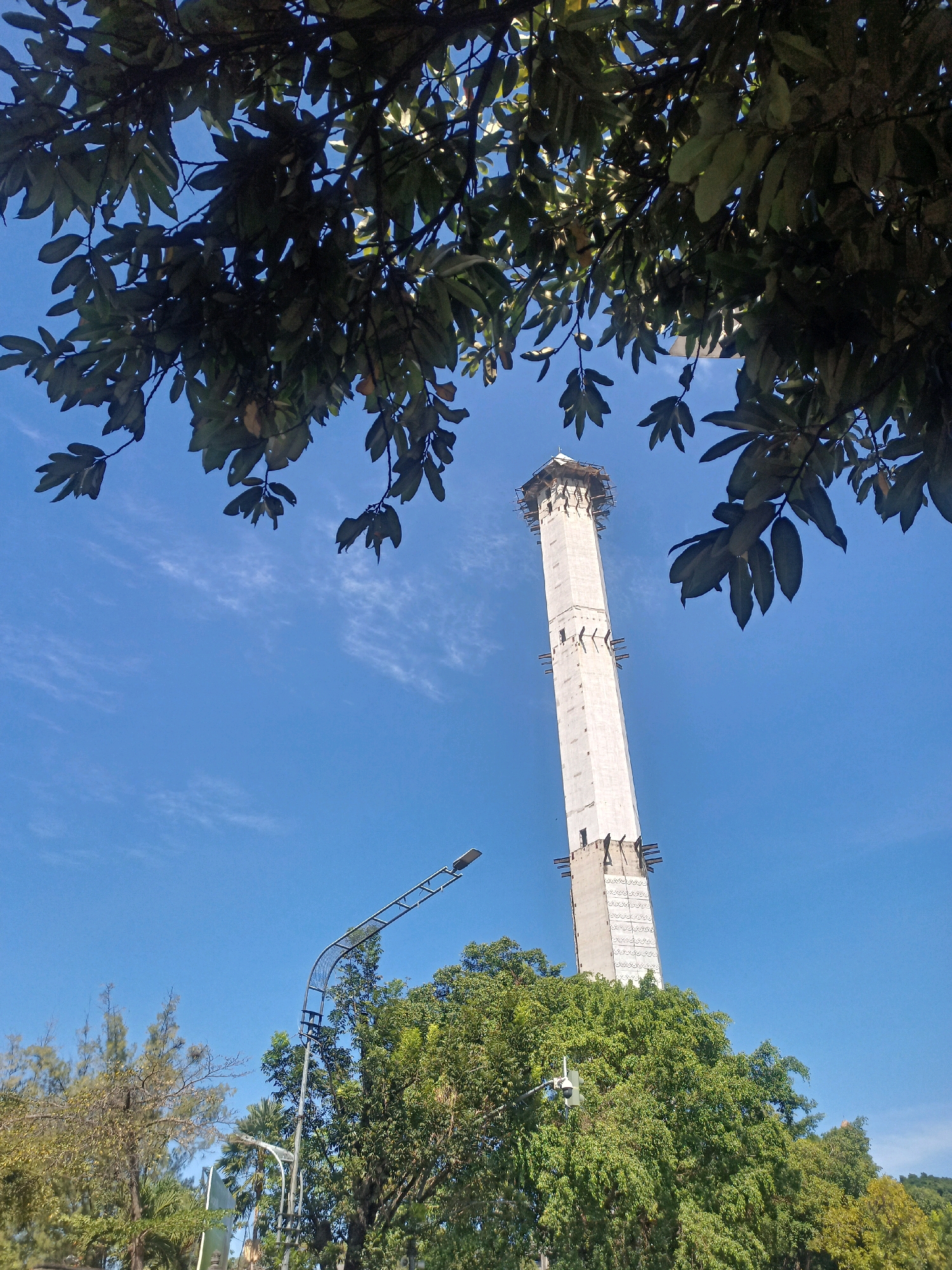 A tall, unfinished concrete tower, possibly under construction, stands against a clear blue sky in Surakarta, Indonesia. The tower is octagonal in shape and appears to be a landmark under development. It is surrounded by lush green trees, and a street lamp is visible in the foreground, suggesting an urban or park setting. The leaves of a tree in the foreground partially obscure the view of the tower and sky. The scene is captured on a bright, sunny day.