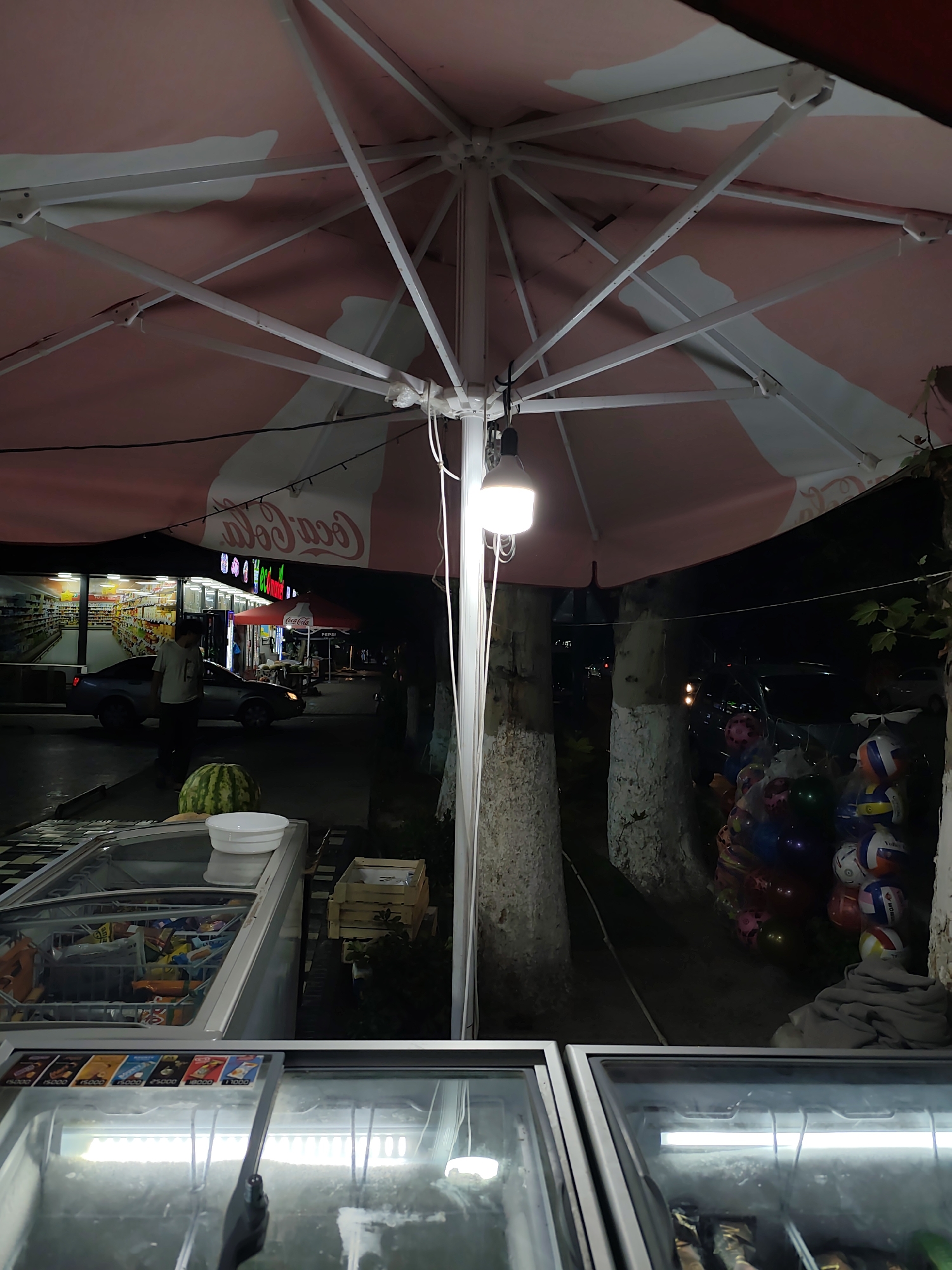 This is a ground-level, eye-level, medium shot photograph taken at night from an outdoor perspective. The scene appears to be a street-level vendor stall or an outdoor section of a shop in Fergana City, Uzbekistan, under a large, pink and white striped umbrella.
The foreground is dominated by two open, chest-style freezers. The left freezer is filled with packaged ice cream and frozen treats, with visible labels and colorful packaging. The right freezer is also illuminated and appears to contain frozen goods, though less detail is discernible.
In the middle ground, a watermelon sits on top of the left freezer, next to a white bowl. To the left of the watermelon, a person is standing, silhouetted against the light from the shop behind them. They are wearing a light-colored t-shirt and dark pants. A gray sedan is parked nearby, facing towards the shop. Behind the car, a brightly lit storefront is visible, displaying rows of goods on shelves. The storefront has prominent signage, including what appears to be "Coca-Cola" in large letters on the umbrella.
To the right of the middle ground, there are two trees with rough, light-colored bark. Stacked bags of balloons and what appear to be volleyballs are visible near the trees, along with a folded gray cloth. A string of lights is visible hanging from the umbrella structure.
The lighting is artificial, with a bright lamp attached to the umbrella illuminating the immediate area. The overall impression is that of a small business operating in the evening, likely selling cold items like ice cream.