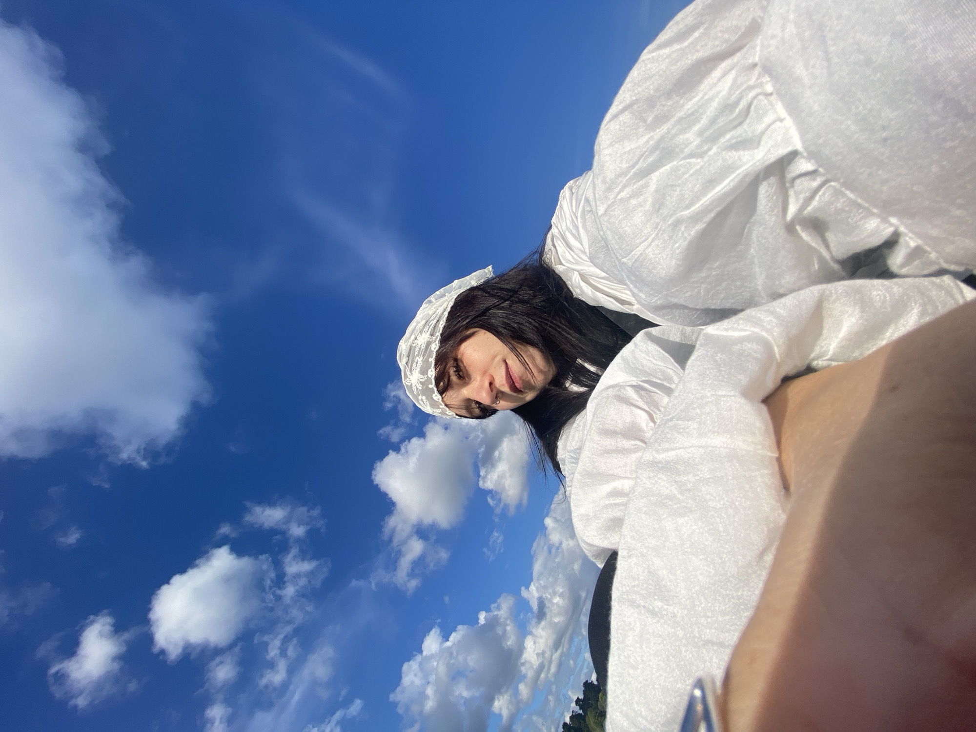 A low-angle, wide shot captures a young woman with dark hair wearing a white lace head covering and a white ruffled garment. She is looking towards the camera, with part of her face obscured by her hair. The background is a vibrant blue sky filled with white, fluffy clouds. The lower portion of the image is dominated by the folds of the white garment and a person's arm and hand, with a silver ring visible on one finger. The setting appears to be outdoors, possibly on a cloudy day.