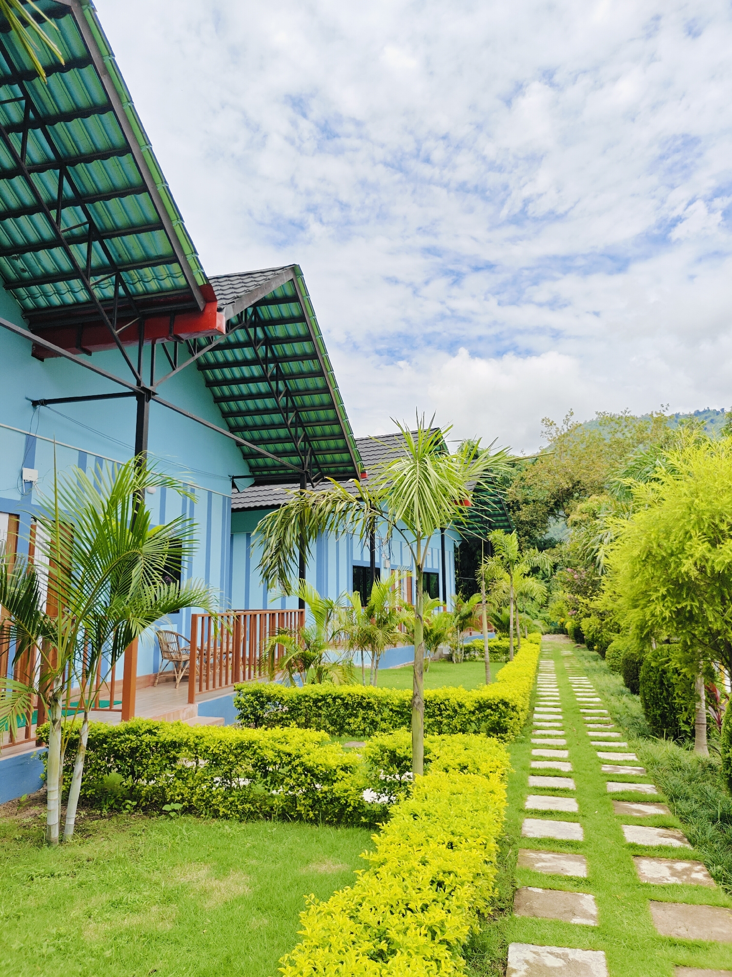 The image depicts a pathway made of stepping stones leading through a lush green garden towards a light blue building with a green corrugated metal roof. The building has a covered porch with wooden railings and is flanked by palm trees and manicured hedges. In the background, a dense forest of trees and hills can be seen under a cloudy sky. The scene is set in the city of Ichchhakamana, Nepal, and appears to be a serene, well-maintained outdoor space.