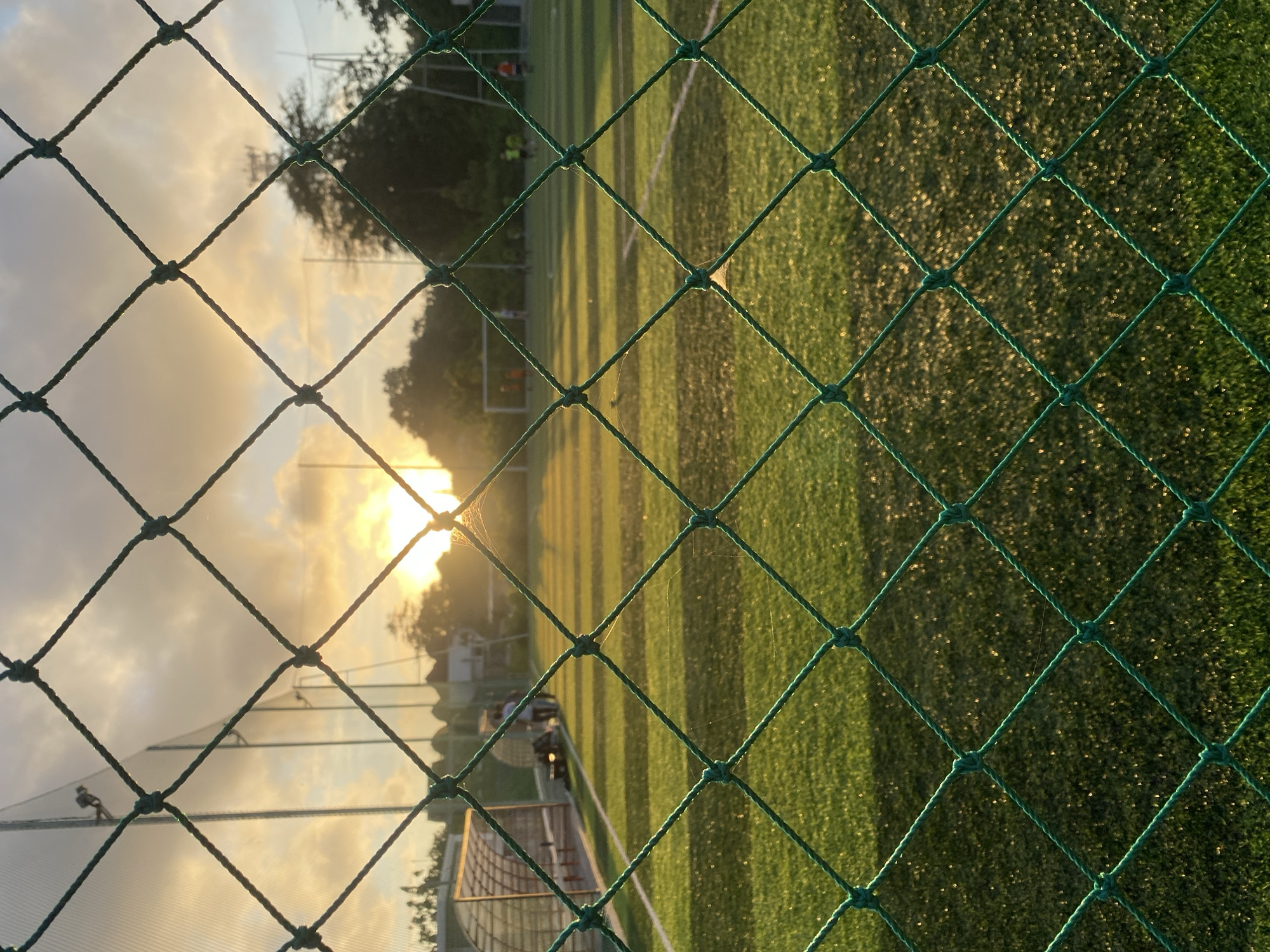 Captured through a green net, the image reveals a sports field under a sky tinged with hues of sunset or sunrise. The artificial turf of the field is marked with white lines. A goalpost stands in the distance. The lighting suggests either dawn or dusk, casting shadows across the field. The location is indicated to be Kuta Selatan, Indonesia.