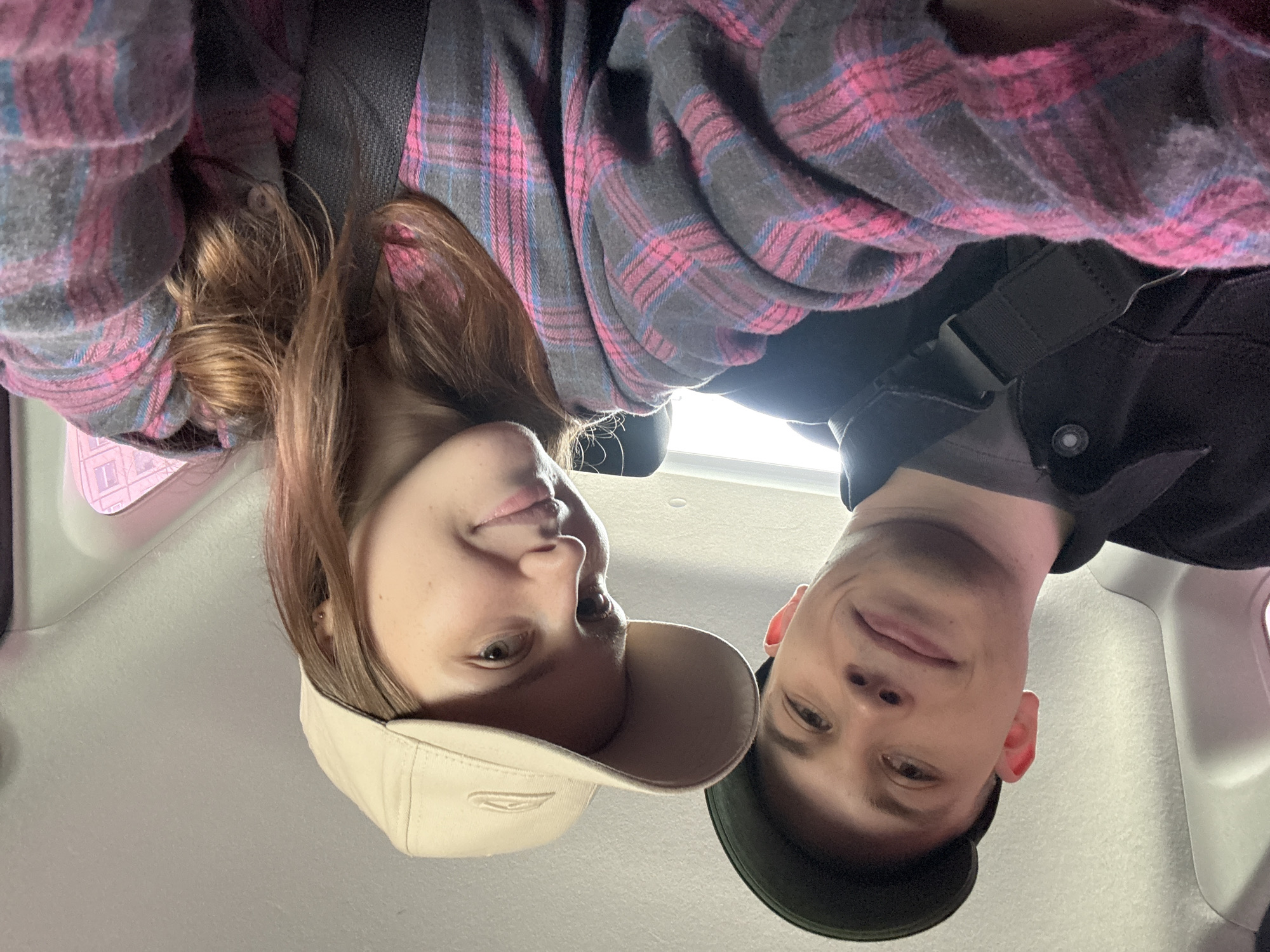 Two young adults, a male and a female, are taking a selfie inside a vehicle, likely a car, based on the interior visible. The male, on the left, is wearing a dark jacket over a gray t-shirt and a dark green baseball cap. The female, on the right, is wearing a pink and gray plaid shirt and a beige baseball cap. Both are looking at the camera and appear to be smiling. The interior of the vehicle shows a light-colored headliner and a hint of a window with pinkish light reflecting from outside. The context of Saint Petersburg, Russia is not discernible from the visual information provided in the image.