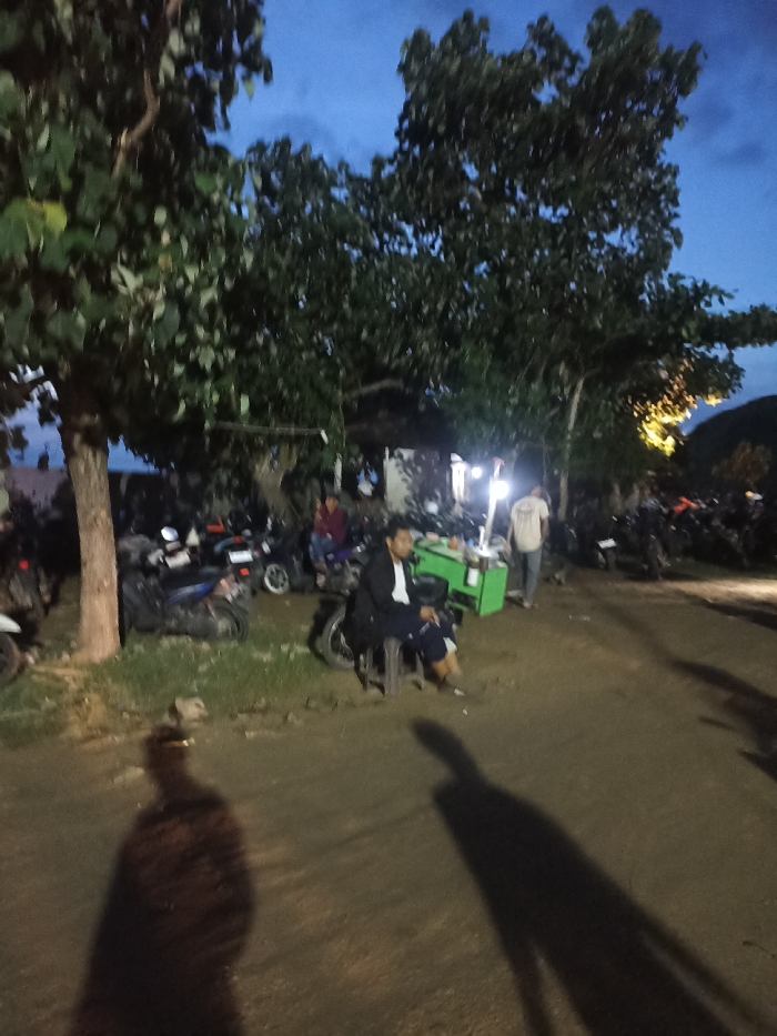 The image captures an outdoor scene at dusk or early night in Kecamatan Pujut, Indonesia. The sky is a deep twilight blue, transitioning to darkness, and artificial lights illuminate key areas.
The scene features several individuals and numerous motorcycles parked on an unpaved, dirt ground. In the mid-ground, a man with dark hair, wearing a dark jacket over a white shirt and dark pants, sits casually on a low stool next to a motorcycle. He is facing to the left, appearing relaxed. Beside him, to his right, is a bright green mobile cart or stall, possibly for vending food or drinks, with various items on its surface. Another person in a light-colored shirt is visible walking away from the camera, near this green cart. Further in the background, additional people are scattered, some near parked motorcycles or illuminated structures.
Large trees with dense foliage frame the upper and left parts of the image, obscuring parts of the sky and background. The ground is rough, with patches of grass near the trees. Prominent, elongated dark shadows of at least two people are cast across the foreground and mid-ground, stretching towards the right side of the frame, indicating a light source behind the camera or to the left. The overall atmosphere suggests a casual gathering spot, possibly a roadside market or a local hangout area, with the presence of street food-style carts and many motorcycles indicating a lively, albeit dimly lit, setting. No legible text is visible in the image.