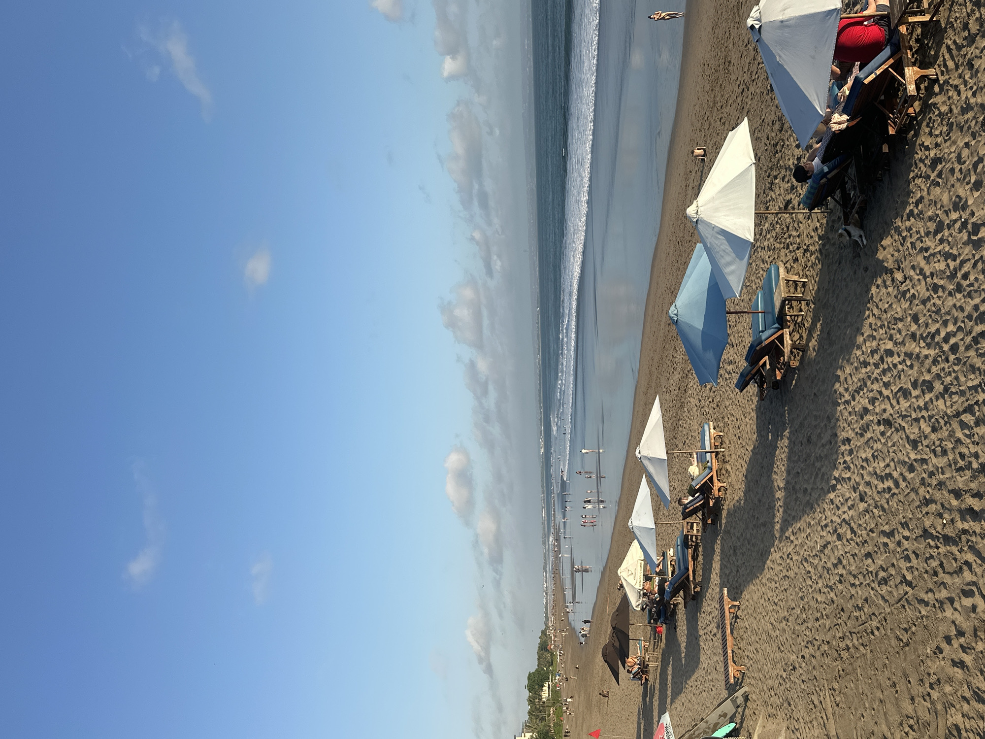 The image depicts a serene beach scene at sunset in North Kuta, Indonesia. The sand is a warm brown, with visible footprints and patterns. Rows of lounge chairs, some occupied by people, are arranged on the sand, shaded by umbrellas of various colors, including white and light blue. Some umbrellas are open, providing shade, while others are closed. A few surfboards lean against a beach umbrella in the foreground.
In the middle ground, the ocean meets the shore with gentle waves creating a foamy white line. People are scattered across the wet sand, some walking, others standing and observing the sea. The water is a deep blue, reflecting the sky. In the distance, a hazy horizon is visible, with faint structures suggesting a town or resort area. The sky is a clear blue, with a few wispy white clouds scattered across it, indicative of a pleasant evening. The overall atmosphere is peaceful and relaxing, characteristic of a popular tourist destination.