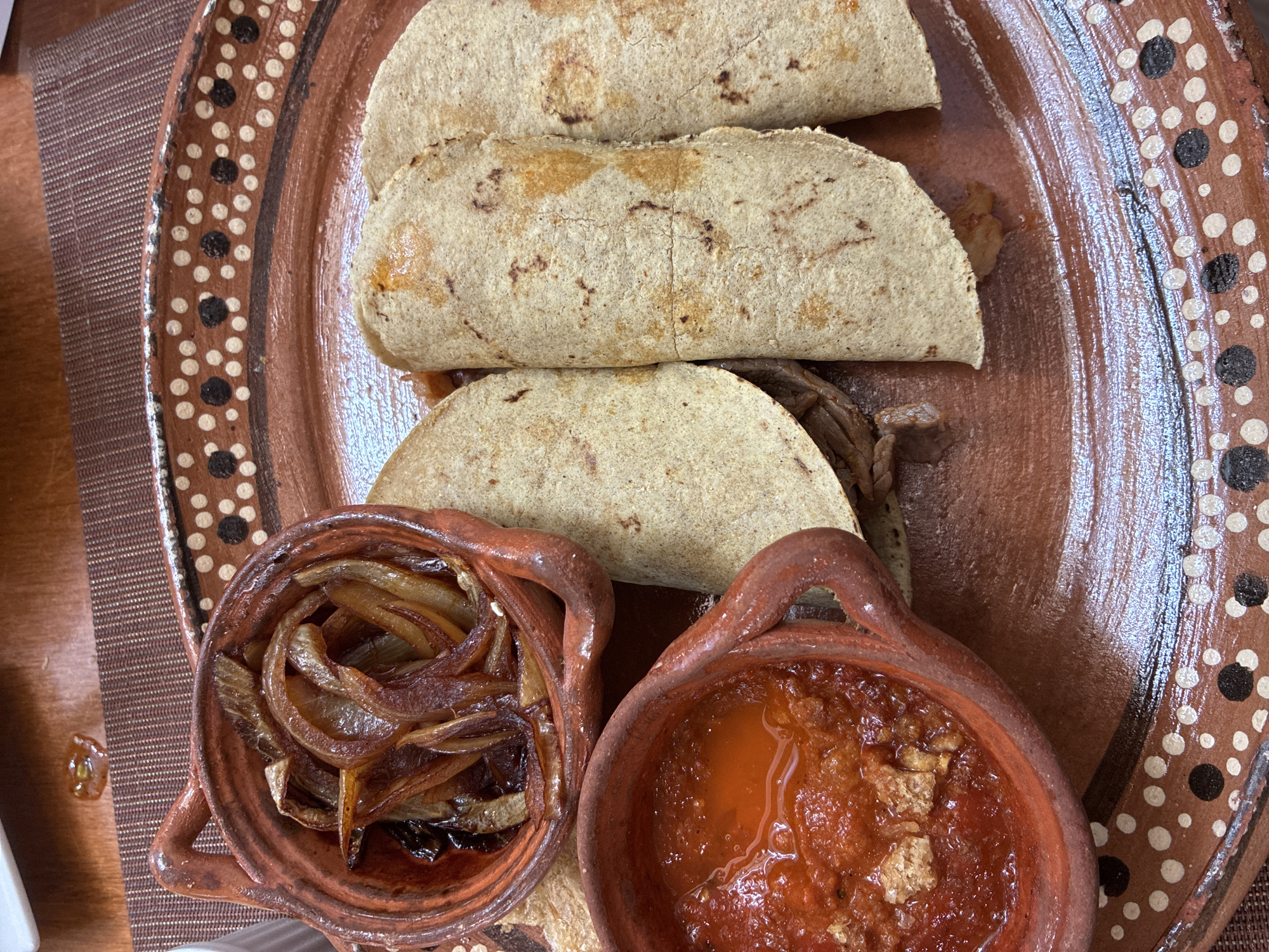 This is a close-up shot of a plate of Mexican food, likely taken in Cuautitlán Izcalli, Mexico. Three tacos, folded in half, sit on a brown, patterned ceramic plate. Next to the tacos are two small clay pots. One pot contains sauteed onions, and the other holds a red salsa. The plate rests on a brown placemat on a wooden table.