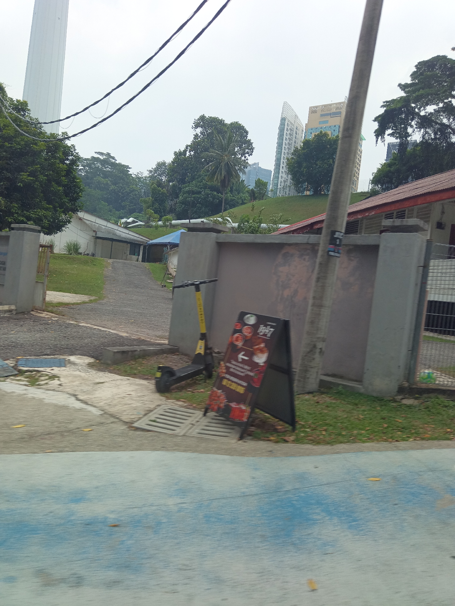 In Kuala Lumpur, Malaysia, an A-frame sign with "1947" written on it stands on a paved pathway. A yellow electric scooter is parked next to the sign. Behind them, a grey wall with a tiled roof building and a fenced area is visible. In the distance, lush green trees and modern buildings, including a tall white tower, are set against an overcast sky. The foreground features a blue painted path.
