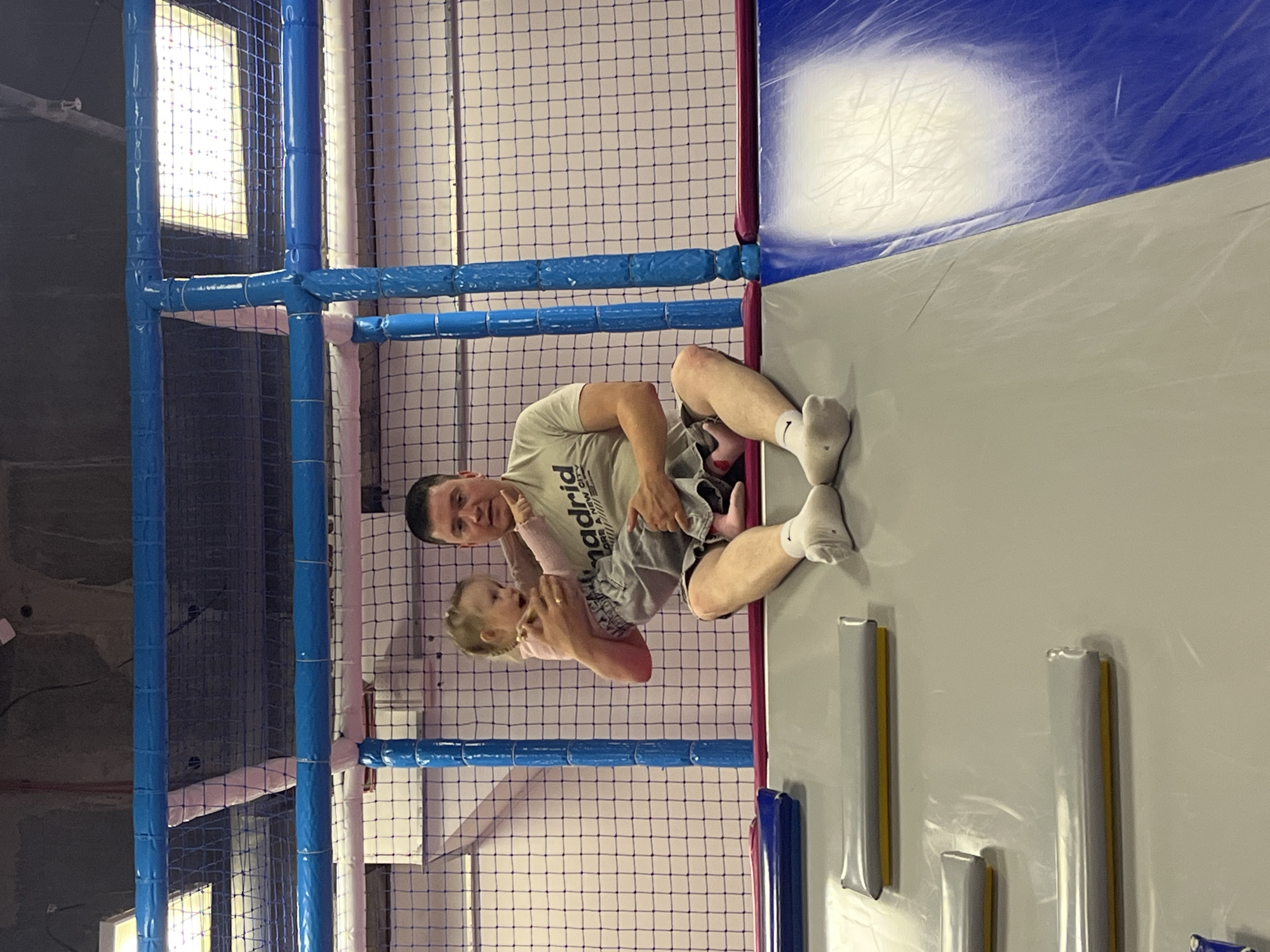 The image captures an indoor recreational facility, specifically a soft play area or trampoline park. An adult male, appearing to be in his late 20s to early 40s with short dark hair, is seated on a grey padded platform. He is wearing a light-colored t-shirt with "Madrid" text, dark shorts, and white socks. He holds a young female child, an infant or toddler, who has light hair styled in pigtails. The child is dressed in a light pink long-sleeved top and grey shorts or skirt, with light pink foot coverings. She is actively pointing her right arm towards the upper left area of the frame, with her mouth open, suggesting vocalization or engagement with something off-camera.
The scene is dominated by safety features typical of such venues. Blue and white padded bars form a structural frame, intertwined with safety netting. The netting behind the individuals exhibits a light pink mesh in certain sections. To the left of the male, several grey rectangular padded elements, each with a yellow underside, are visible on the grey platform, possibly serving as steps or obstacles. To his right, a reflective blue padded surface extends downwards, indicative of a slide or a similar soft play feature. The ceiling above is dark, revealing exposed pipes and rectangular fluorescent light fixtures, characteristic of an industrial or commercial building adapted for recreation. The overall lighting appears artificial and bright.