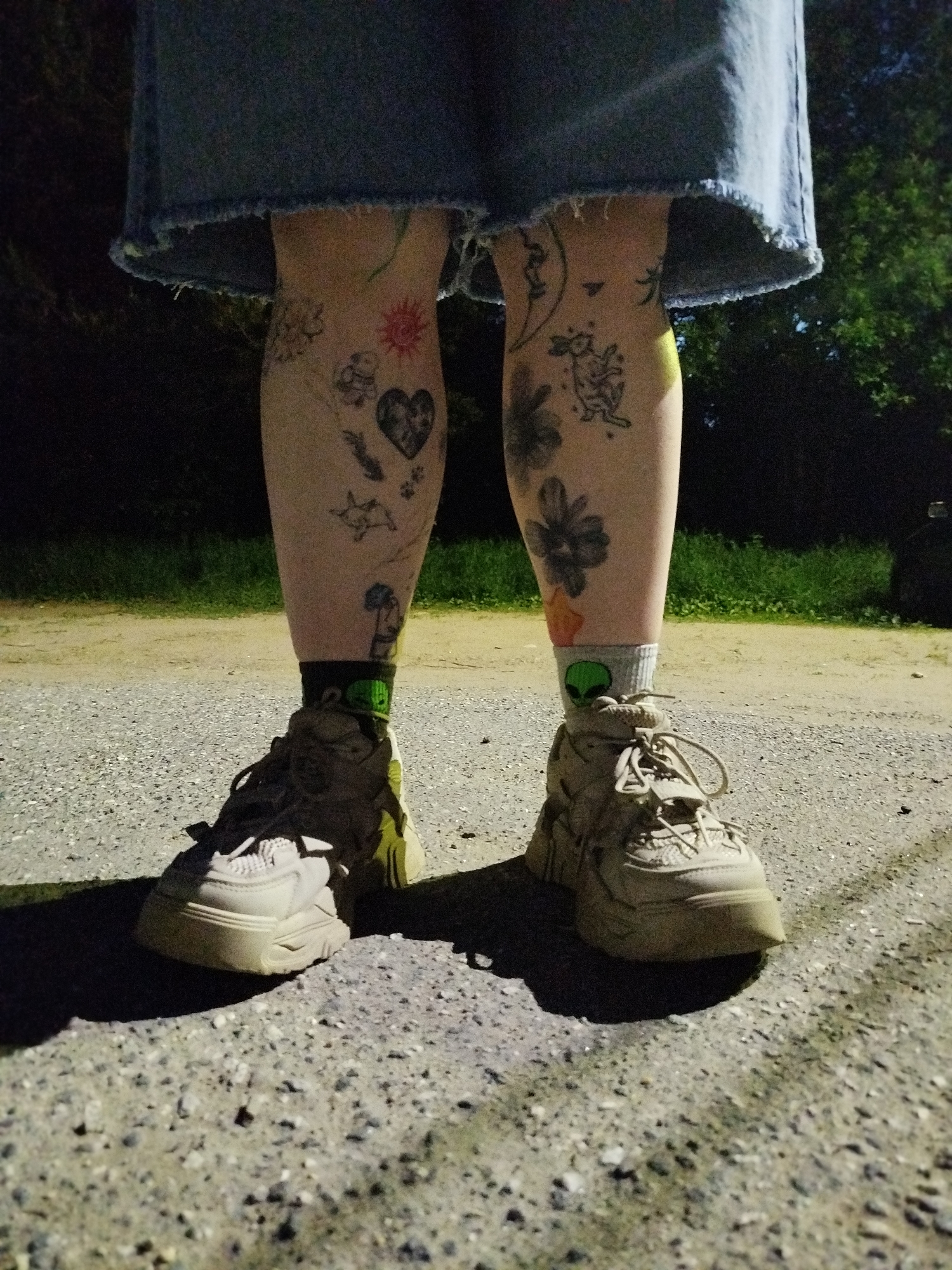 An outdoor, medium shot reveals a person from the knees down, standing on a gravel road or path, location unknown. The person is wearing a short, light-blue denim skirt. The legs are fair-skinned with multiple tattoos, including a heart, a sun, flowers, and a moon. The person is wearing sneakers and socks, one of which has a green alien head. The background consists of dark trees and foliage. The lighting is dim, suggesting it is dusk or nighttime.