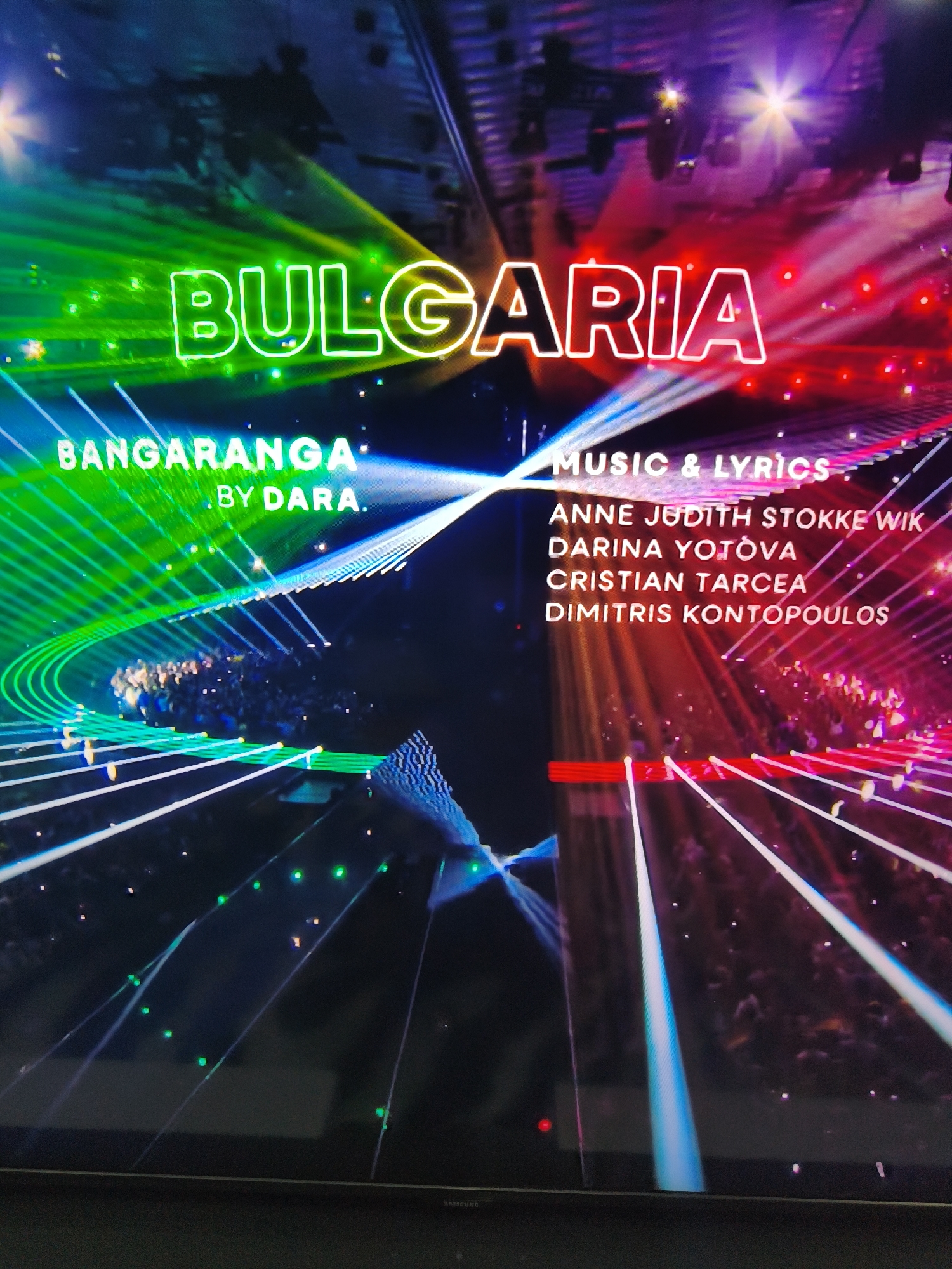 The image displays a television screen showing credits for a musical performance, captured in Svilengrad, Bulgaria. The screen presents a vibrant, high-energy stage setting illuminated with striking green, blue, and red lights and laser beams. A crowd of people is visible on the stage or within the immediate audience area, suggesting a live concert or large-scale event.
Prominently displayed on the screen is the word "BULGARIA" in large, glowing, neon-style letters that transition from green on the left to red on the right. Below this, "BANGARANGA" and "BY DARA." are shown in smaller green text, identifying the song and artist. To the right, the section "MUSIC & LYRICS" is visible, followed by a list of names credited for the music and lyrics: "ANNE JUDITH STOKKE WIK," "DARINA YOTOVA," "CRISTIAN TARCEA," and "DIMITRIS KONTOPOULOS."
The overall scene on the screen depicts a dynamic stage environment with colorful light effects, indicating a modern musical production. The physical television screen, visible at the bottom of the image, has a black bezel and a subtle "SAMSUNG" logo in the bottom right corner. The perspective of the image is slightly angled, suggesting it was taken by a person watching the television.