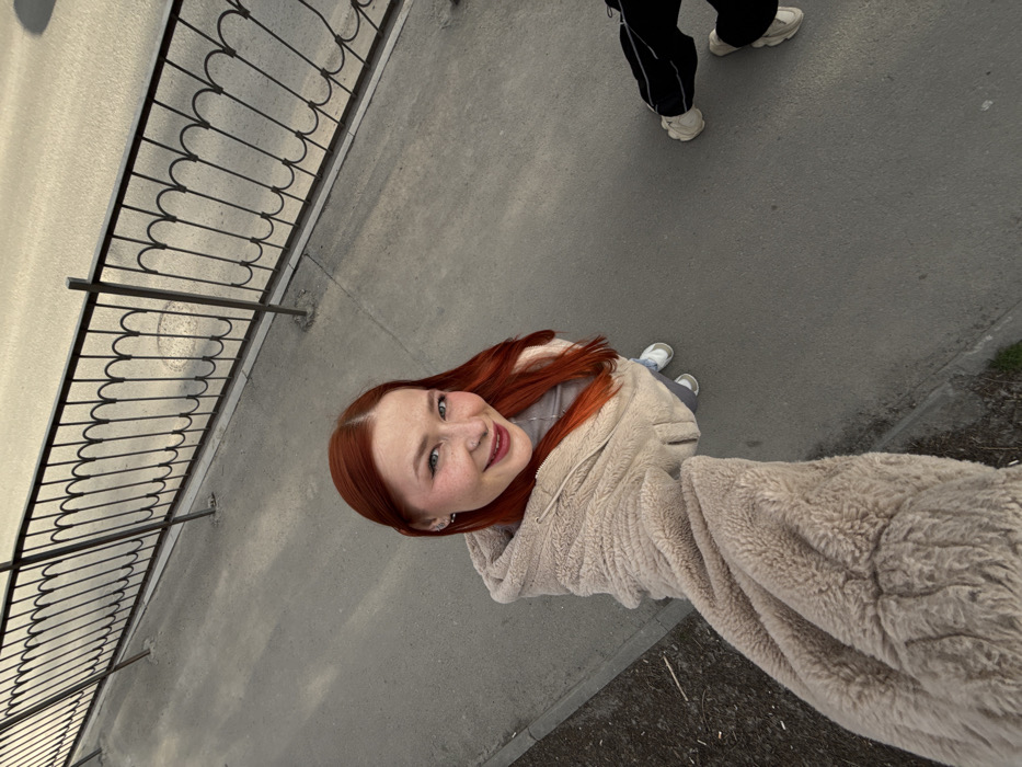 A high-angle outdoor shot depicts a young female and the lower extremities of a second individual on a grey paved surface in Rostov-on-Don, Russia. The primary subject, positioned centrally, has long, vibrant red hair, a fair complexion, and light-colored eyes. She is smiling directly at the camera, displaying a small stud nose piercing on her right nostril and small hoop earrings. She wears a light beige, textured hooded jacket over a gray top, light-colored pants, and white sneakers. Her right arm is extended forward, holding the camera for a selfie. To her right, the lower legs and feet of another person are visible, clad in dark pants with a white lateral stripe and light-colored sneakers. A black metal fence with repeating decorative arch patterns runs horizontally across the upper frame, separating the paved area from a lighter grey road surface. A circular manhole cover is visible on the road behind the fence. The scene is illuminated by natural daylight.