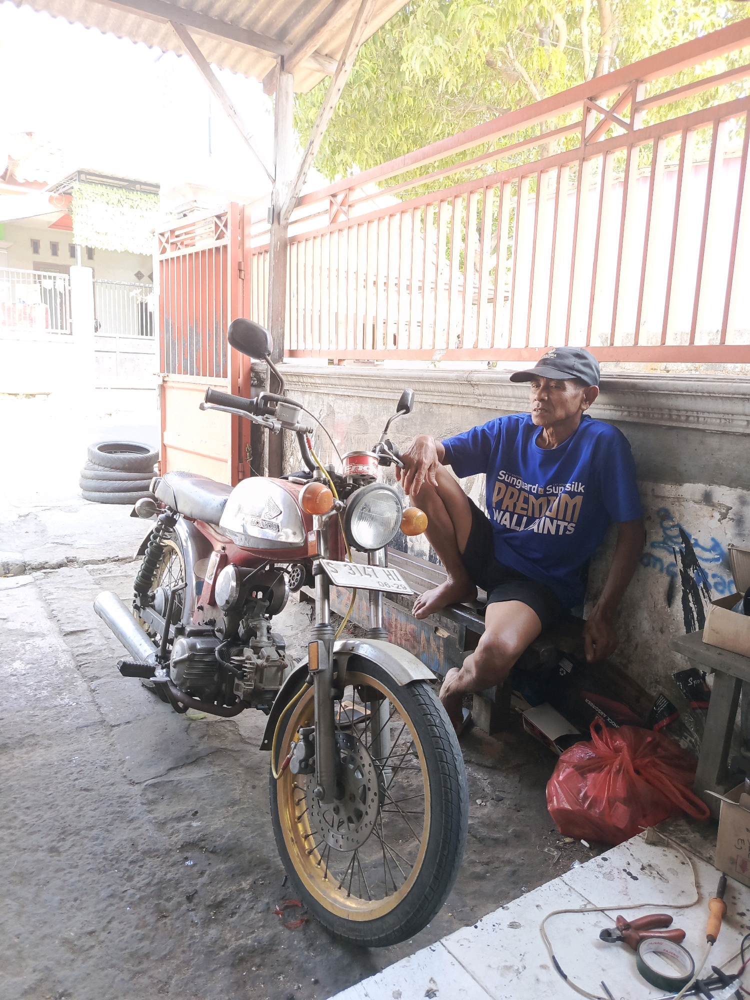 A man wearing a blue t-shirt and a dark baseball cap sits on a wooden bench next to a red vintage motorcycle. The man is looking to his left and has his left arm resting on the motorcycle. The motorcycle is parked on a concrete floor, and there are stacks of tires to the left of the motorcycle. The scene appears to be outdoors, possibly in a workshop or garage, with a corrugated metal roof overhead and a red metal fence in the background. The environment is somewhat cluttered with various objects, including tools and a red plastic bag. The location is identified as Semanding, Indonesia.