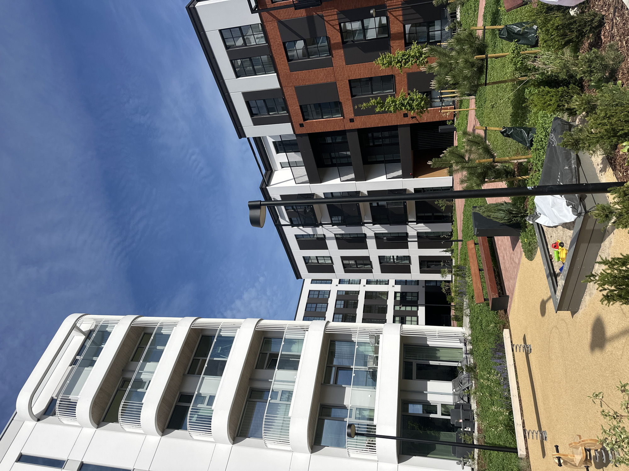 This is an eye-level, medium shot of a modern apartment complex in Tallinn, Estonia, on a sunny day with a partly cloudy blue sky. The buildings feature a mix of white, black, and red brick facades, with contemporary balconies and large windows. In the foreground, a children's play area is visible, complete with a sandbox, a see-saw, and a spring rider. The courtyard is landscaped with grass, small trees, and shrubs, and includes a wooden bench. No people are present in the image.