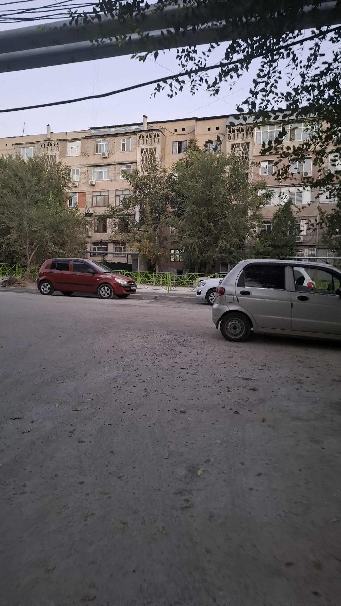The image depicts an outdoor scene in the city of Shymkent, Kazakhstan, likely a residential area with an apartment building and a street. In the foreground, the rough, grey asphalt of a road is visible, with scattered debris like leaves and small stones. To the left, a red hatchback car is parked on the side of the road. Further down the street, a silver hatchback car is also parked, with a white car partially visible behind it.
A green metal fence runs along the edge of the sidewalk, separating the street from a grassy area and trees. Behind the fence, several trees with green foliage stand tall. In the background, a multi-story apartment building dominates the scene. The building has a light-colored facade and features numerous windows, some with balconies or decorative elements. The sky above is overcast, suggesting a cloudy day. The overall impression is a typical urban streetscape in a residential district.