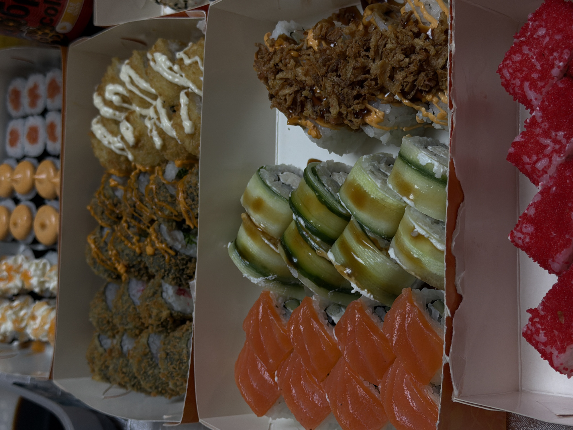 The image displays a close-up, elevated view of an assortment of sushi and sushi rolls arranged in several open cardboard takeout boxes. The scene suggests a large order ready for consumption.
From left to right, the boxes reveal a variety of sushi preparations:
* The leftmost box contains two rows of what appear to be maki rolls with white rice and orange/red centers, followed by rolls topped with an orange sauce, then tempura-fried rolls drizzled with a white sauce, and finally, more tempura-fried rolls with an orange sauce.
* The middle box showcases three distinct types of rolls: the top row features rolls heavily topped with crispy fried onions and drizzled with a dark sauce; the middle row consists of rolls elegantly wrapped in thin slices of green cucumber; and the bottom row presents rolls topped with vibrant, thinly sliced salmon.
* The rightmost box, partially visible, contains rolls generously coated in bright red roe or tobiko, giving them a distinct texture and color.
The packaging indicates these are takeout or delivery items. A red can, possibly a soft drink, with visible text "CO" (likely part of "COLA") is partially seen in the top-left corner, leaning over one of the boxes. The overall impression is one of a diverse and colorful sushi platter. No people are visible, and the setting appears to be an indoor environment.