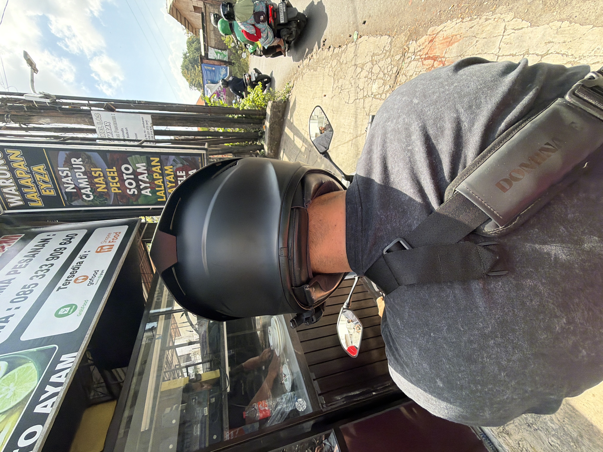 This is a first-person perspective, eye-level, outdoor, daytime, full-body shot of a person wearing a black helmet and a grey t-shirt, seated on a motorcycle in Kuta Selatan, Indonesia. The rider's back is facing the camera, and they are wearing a black sling bag. The background shows a street with a warung (food stall) displaying a menu of local dishes like "Nasi Campur," "Nasi Pecel," and "Soto Ayam." The warung also advertises delivery services through GoFood, GrabFood, and Shopee Food, with a phone number for orders. A woman and two children on another motorcycle are visible further down the street, also heading in the same direction. The environment appears to be a typical street scene in a tropical urban area, with a paved road, a sidewalk, and vegetation. The weather is sunny, with a clear blue sky and some clouds. The reflection in the motorcycle's side mirror shows two people in the rider's vicinity.