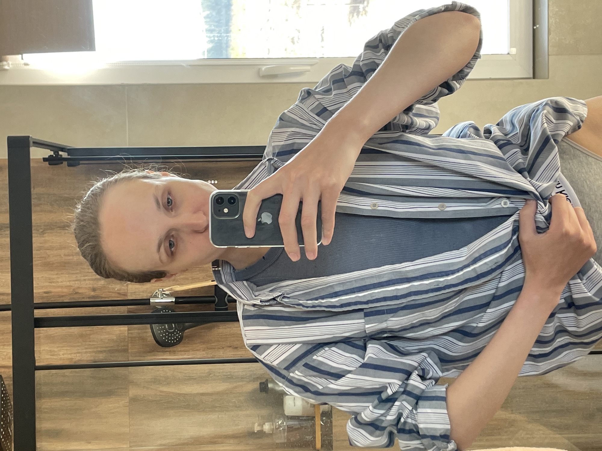 A person is taking a selfie in a mirror. They are holding a phone in their left hand, which is partially covering their face. The person is wearing a striped, button-down shirt over a solid-colored top. The background shows a bathroom setting with a window, shower fixtures, and tiled walls. There's no definitive information in the image to confirm that the location is Подшивалово, Russia.
