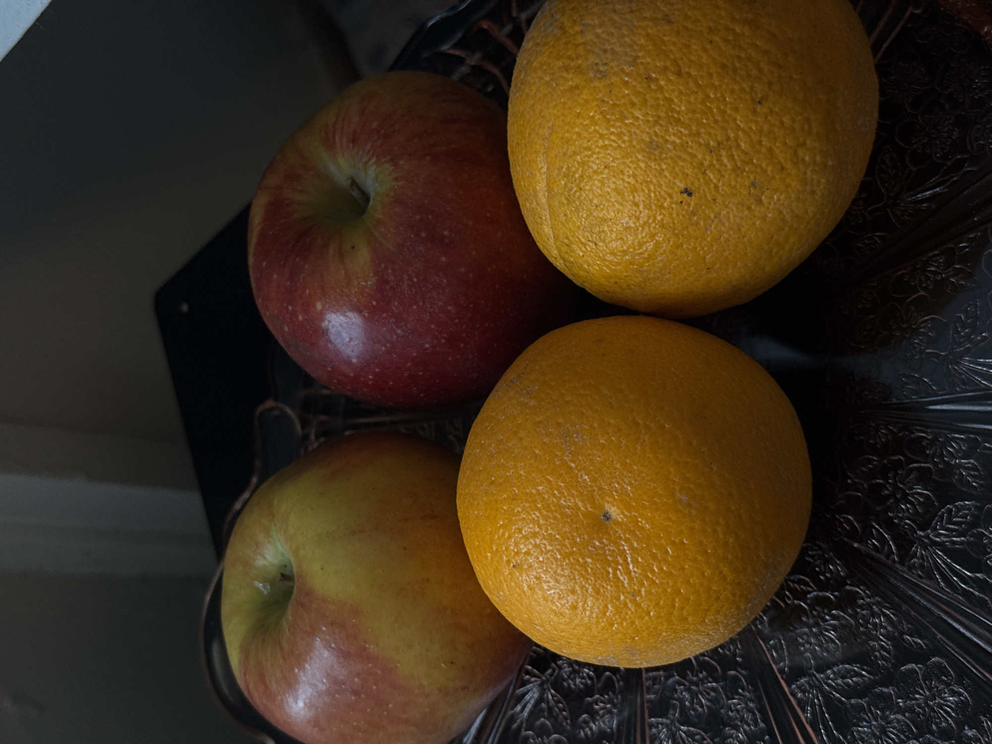 A close-up, vertical shot features four pieces of fruit arranged on a dark, ornate platter. Two apples, one predominantly yellow with red blush and another deep red, are positioned in the upper portion of the frame. Below them sit two oranges, their textured skins clearly visible. The lighting is dim, casting subtle shadows and highlighting the textures of the fruit and the intricate pattern on the platter. The background is dark and out of focus, suggesting an indoor setting. The image does not depict any identifiable landmarks or provide clues to its specific location within Saint Paul, United States of America.