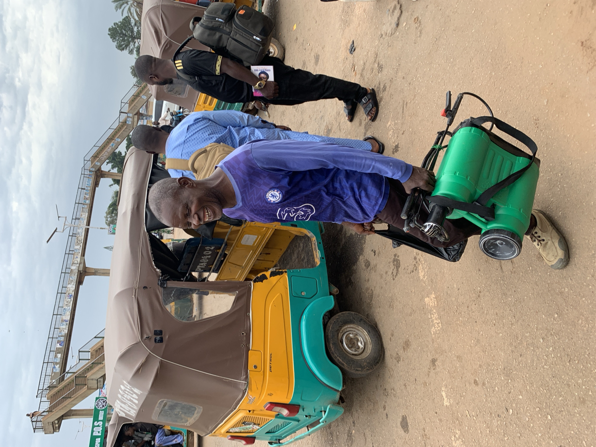 In Gura Top, Nigeria, a man with a wide smile walks on a dusty road, holding a green sprayer. He wears a long-sleeved blue shirt with a white logo and tan shoes. Behind him, a yellow and turquoise auto-rickshaw is parked, its canopy partially obscuring the view of other people and vehicles. Further in the background, a pedestrian bridge with stairs spans overhead, and a sign for "P.O.S Outlet" is visible. The sky is overcast.