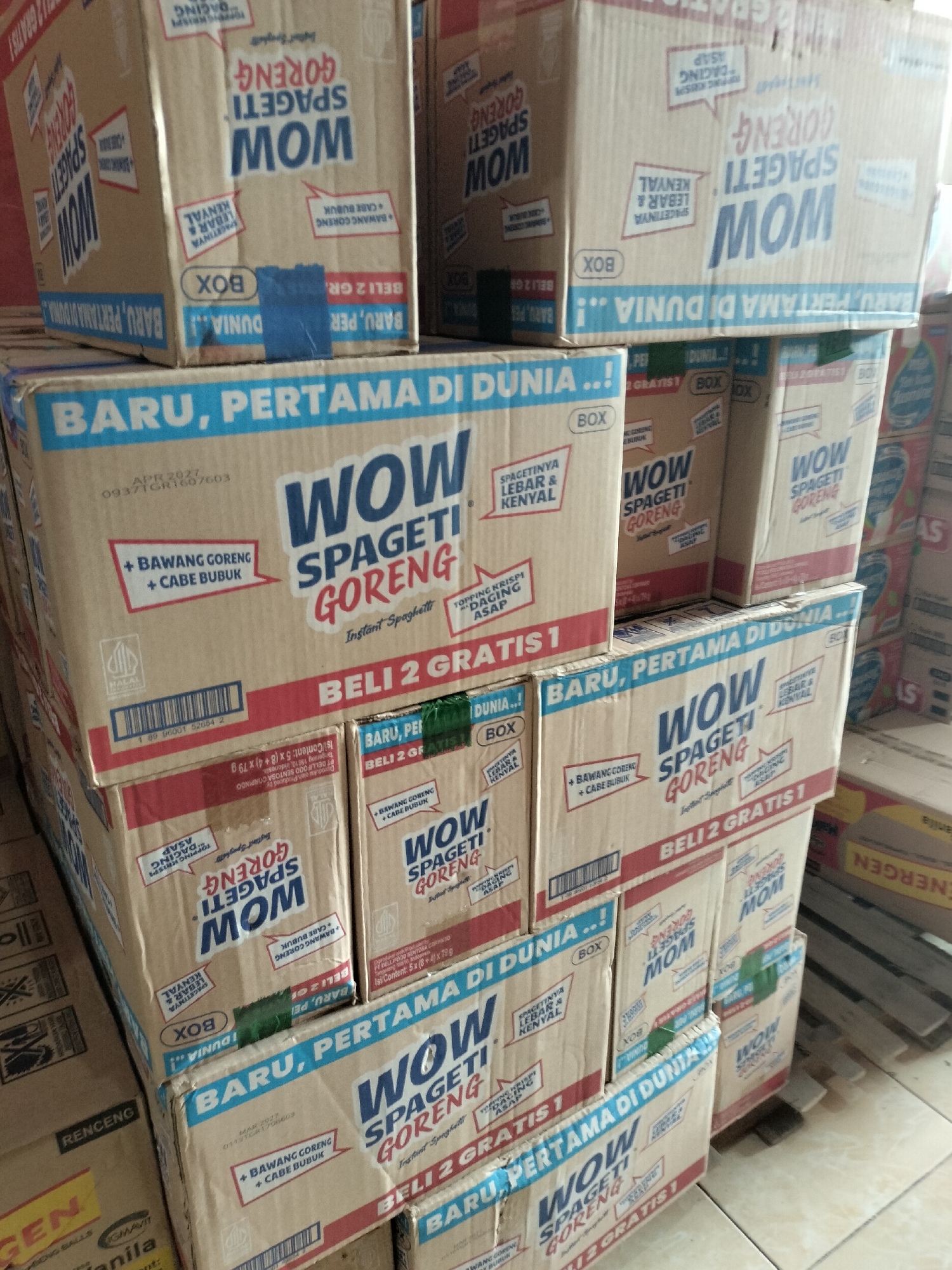 The image shows a stack of cardboard boxes, likely at a retail or storage location in Langsa, Indonesia. The boxes are predominantly brown with blue and red text. They feature branding for "WOW SPAGETI GORENG" and "MOM SPAGETI GORENG". Some boxes indicate "BARU, PERTAMA DI DUNIA!!" (New, First in the World!!) and "BELI 2 GRATIS 1" (Buy 2 Get 1 Free). Various other text, including ingredient lists and barcodes, are visible on the boxes. The environment appears to be an indoor space with a tiled floor. The lighting suggests it could be daytime, with some artificial light also present. The focus is entirely on the product packaging.