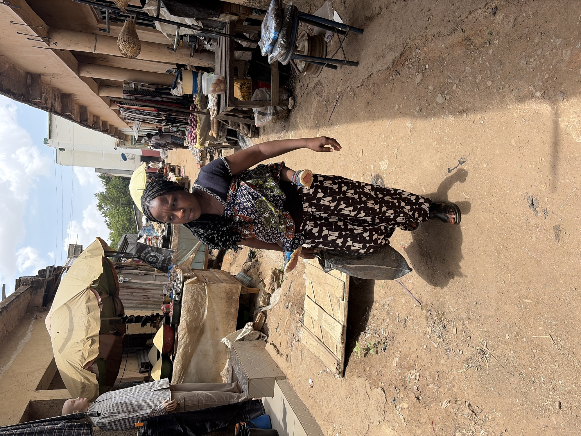 A vibrant outdoor market scene in Jos, Nigeria, is depicted under bright daylight. The main subject is a woman positioned slightly off-center in the foreground, facing the viewer with a slight smile. She has dark braided hair and is dressed in a dark top, a colorful patterned fabric wrapped around her midsection, and a long skirt featuring a black and white abstract pattern. A light-colored bracelet is visible on her right wrist, and she appears to be carrying a dark bag or package at her hip.
The bustling street is lined with market stalls and modest multi-story buildings. Various goods are displayed along the pathway, including clusters of red onions, packaged items, and other merchandise. Baskets hang from overhead structures. To the left, a large, light-colored umbrella provides shade next to a male mannequin dressed in a plaid shirt and trousers. The ground is unpaved, dusty, and uneven, littered with small stones and dry plant matter.
In the background, other individuals are visible, contributing to the sense of market activity. The sky above is blue with scattered white clouds, indicating sunny weather. Shadows cast on the ground suggest it is daytime, likely midday or early afternoon. A black bag hanging in the background bears the text "SHOPPING" and an "S" logo, reinforcing the commercial environment.