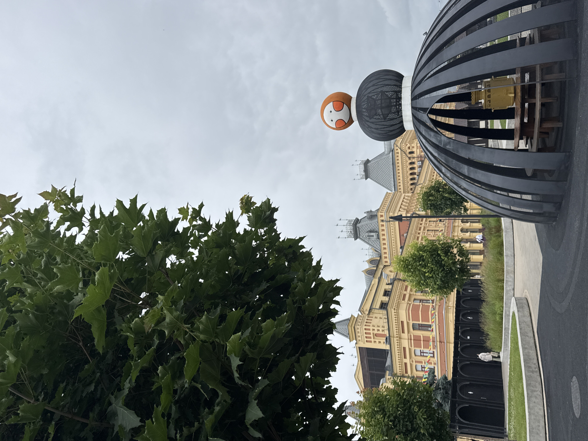 In Nizhny Novgorod, Russia, a street scene unfolds under an overcast sky. In the foreground, a large tree with dark green leaves partially obscures the view. To the right, a modern black metal gazebo-like structure with a rounded top and open sides shelters a wooden picnic table and benches. Perched atop the structure is a large, round, orange object with a cartoonish face, featuring two white circles with orange outlines for eyes and a small orange mouth. Behind the gazebo, a yellow building with red accents and ornate architectural details stands prominently. In front of the building, a row of black arched entrances with signs resembling restroom symbols lines a fenced area. A person in a light-colored coat walks towards the entrances. Green grass and small trees are visible between the gazebo and the building. The ground in the foreground is paved with dark asphalt.