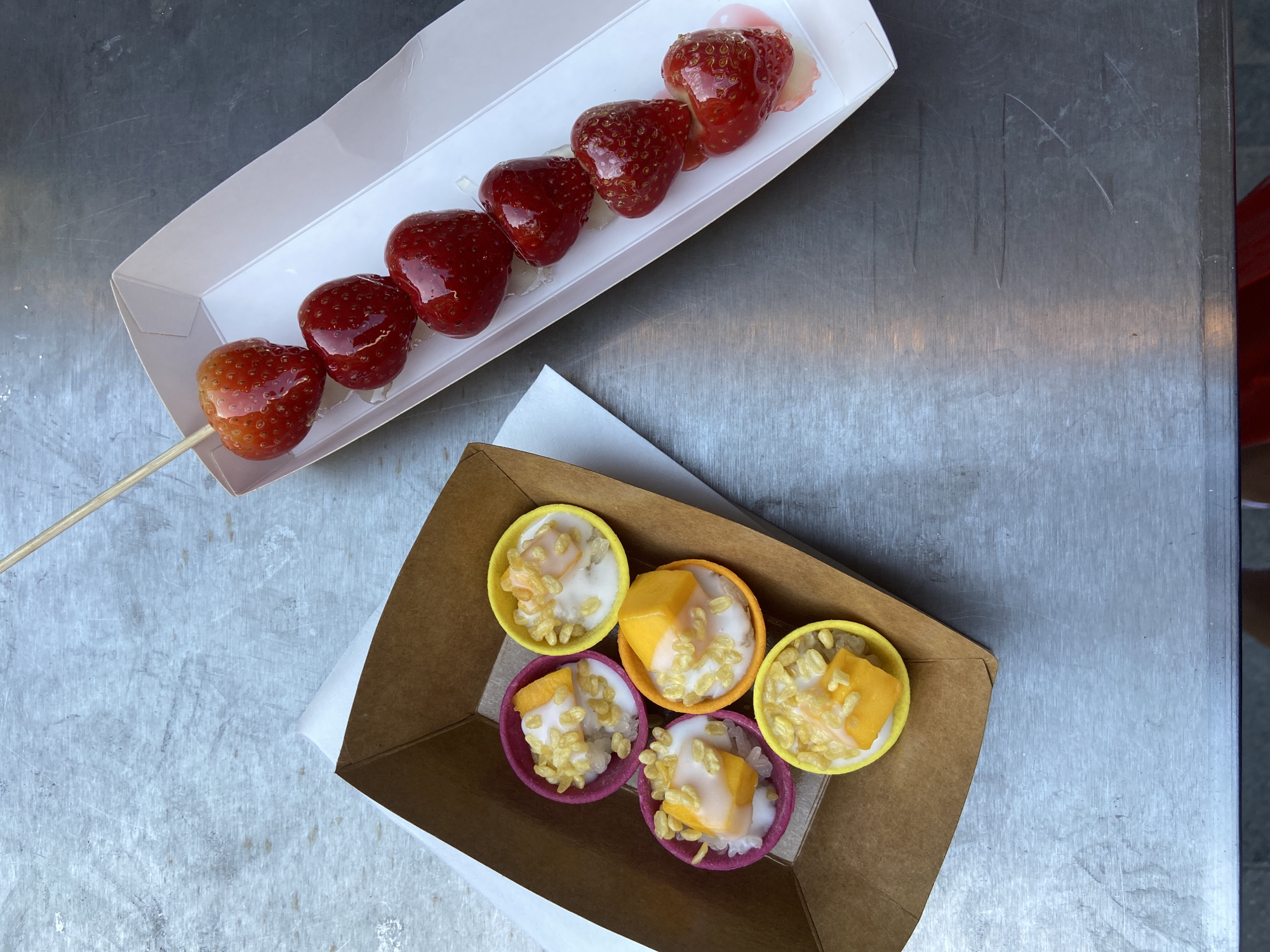 The image shows two food containers sitting on a metal surface. One container holds a skewer with six strawberries, all covered in a clear, shiny coating. The other container, which is placed on a white napkin, holds five small cups filled with what appears to be a rice dish topped with mango. The cups are different colors, yellow, orange, and purple. It appears to be a street food scene, but the location in the city of London, United Kingdom, cannot be definitively determined from the given image.