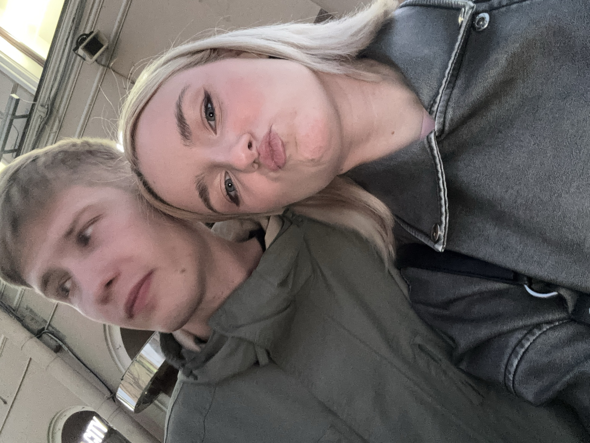 Two individuals, a male and a female, are captured in a close-up, low-angle selfie, with the male's head and upper body on the left and the female's face in the foreground on the right. The female is making a kissing face, with her lips pursed. Both individuals are wearing jackets, with the male in a green hooded jacket and the female in a dark grey jacket with visible stitching and metal snap closures. They appear to be outdoors, possibly in a city setting, with architectural elements visible in the background. A sign with the word "CITY" is partially visible on the left. The lighting suggests it is either dusk or an indoor environment with artificial lighting. The exact location in Saint Petersburg, Russia, cannot be determined from the image alone.