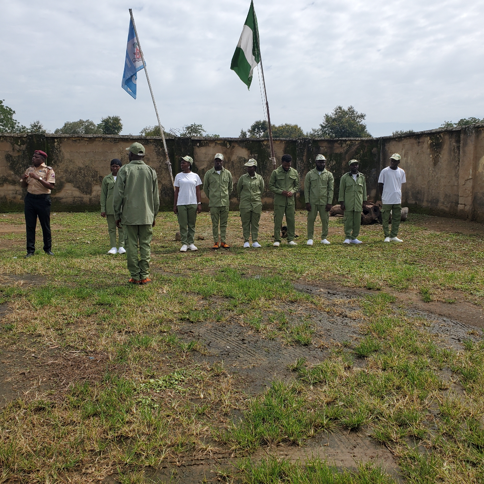 This media file captures an outdoor assembly in Toro, Nigeria, under a cloudy sky, likely in the late morning or early afternoon. The scene is set in a grassy, somewhat uneven area, bordered by a textured concrete wall. Two flagpoles stand prominently. On the left, a blue flag with white symbols and what appears to be a red emblem is raised. To its right, a Nigerian flag with green and white stripes flutters.
The primary subjects are a group of individuals, most of whom are dressed in identical olive-green uniforms and caps. They are lined up in what appears to be an organized formation. A single individual on the far left stands out, wearing a tan uniform with a red beret, seemingly addressing the group or overseeing the assembly. Among the uniformed individuals, at least two are women, distinguished by their uniforms and hairstyles. One woman on the far right of the uniformed group is wearing a white t-shirt and olive-green trousers, but without the uniform shirt.
The setting suggests a public space or an institution, possibly a training ground or a ceremonial area, given the flags and the organized assembly. The ground is covered with patchy grass and some muddy or wet patches, indicating recent rainfall or damp conditions. The overall mood appears formal and attentive. There is no legible text visible on the uniforms or any other objects in the frame.
