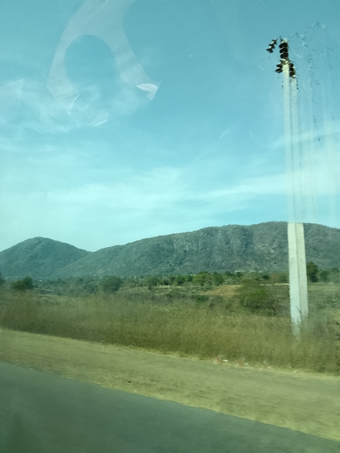 This image depicts an outdoor landscape scene in Jibam, Nigeria. The foreground features a dark, paved road extending diagonally from the lower left towards the center, alongside a strip of light brown, dry earth. The midground consists of extensive fields of dry, yellowish-brown grasses and scrubland, dotted with scattered green trees and bushes. In the background, two prominent, dark green, tree-covered hills or mountains rise against a clear, light blue sky with minimal cloud cover. A tall, slender, light-colored utility pole is positioned on the far right, topped with dark utility components. Reflections, including an inverted U-shape and irregular light patterns, are visible across the transparent surface through which the photograph was taken, predominantly in the upper left, along with vertical streaks on the right side. No individuals, animals, or discernible actions are present within the scene.