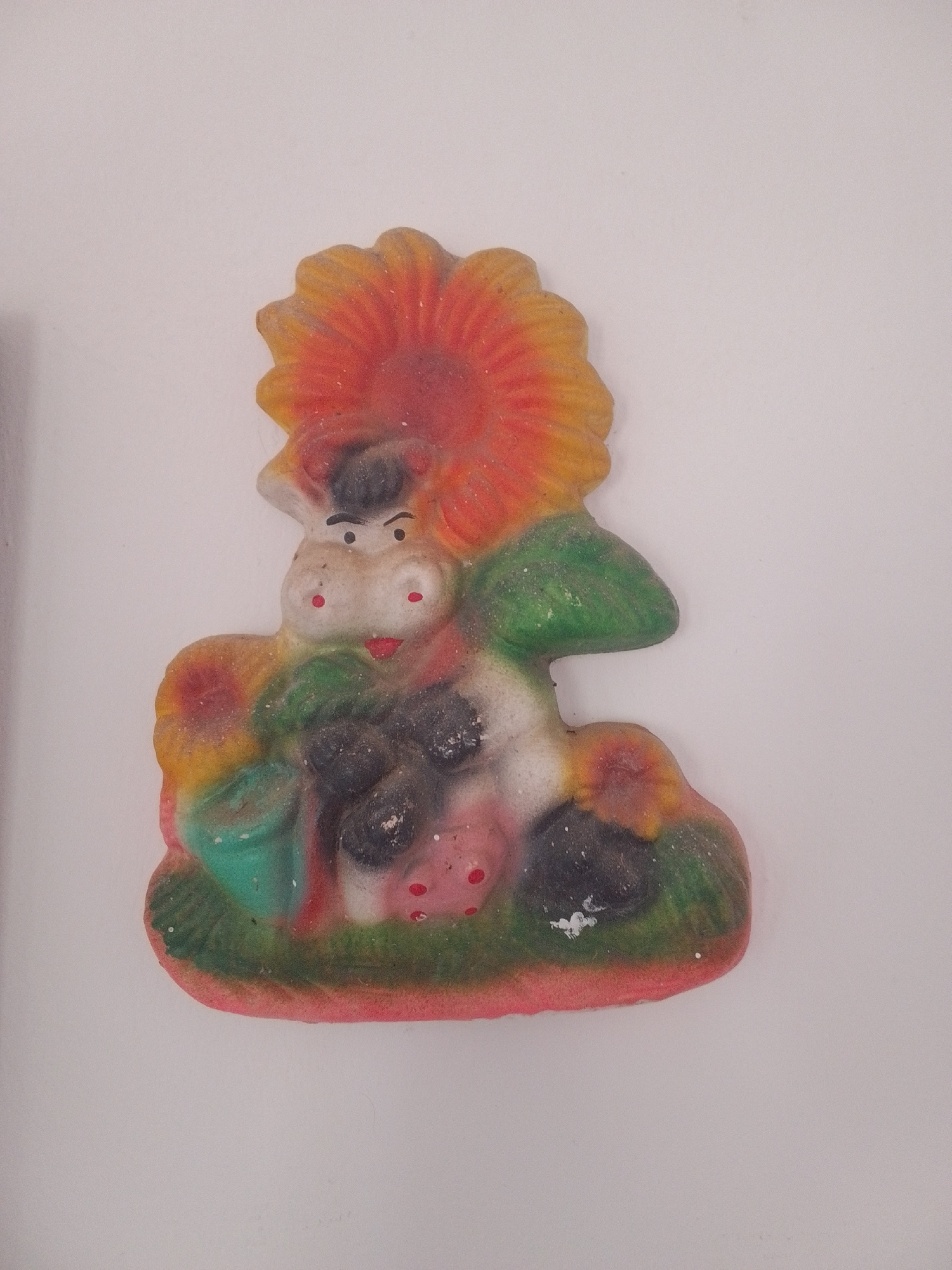 This is a colorful, three-dimensional ceramic figurine, possibly a decoration or a toy, with a white background. The figurine depicts a stylized, cartoonish cow seated among a large, red and yellow sunflower and green foliage. The cow has a white face with black eyebrows, two red dots for cheeks, and a smiling red mouth. Its body is black and white, with black patches. The cow is holding a small green bucket in its left hand. To the right of the cow is a green mushroom shape. The base of the figurine is green, resembling grass, and has a red border. The overall style is whimsical and playful. There are no visible text or inscriptions on the figurine. The scene appears to be a simple, cheerful, and possibly rural or garden-themed setting. The lighting is even, suggesting it was photographed indoors.
