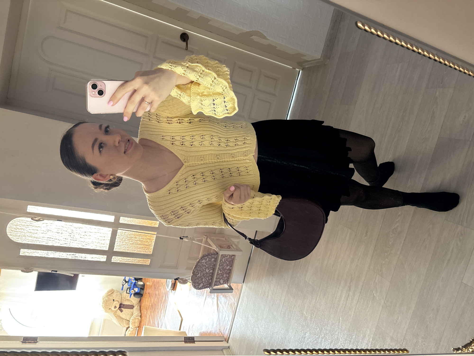 A young woman is taking a selfie in a mirror. She is wearing a yellow crocheted cardigan, a black skirt, and black tights with black socks. She has a brown purse hanging off her shoulder. The background shows the interior of a room with a white door, light-colored flooring, and a glimpse into another room with a television, teddy bear, and child's toys. The mirror frame is gold. The location is indeterminate, and there is no information to confirm that the image was taken in the city of Ферма, Russia.