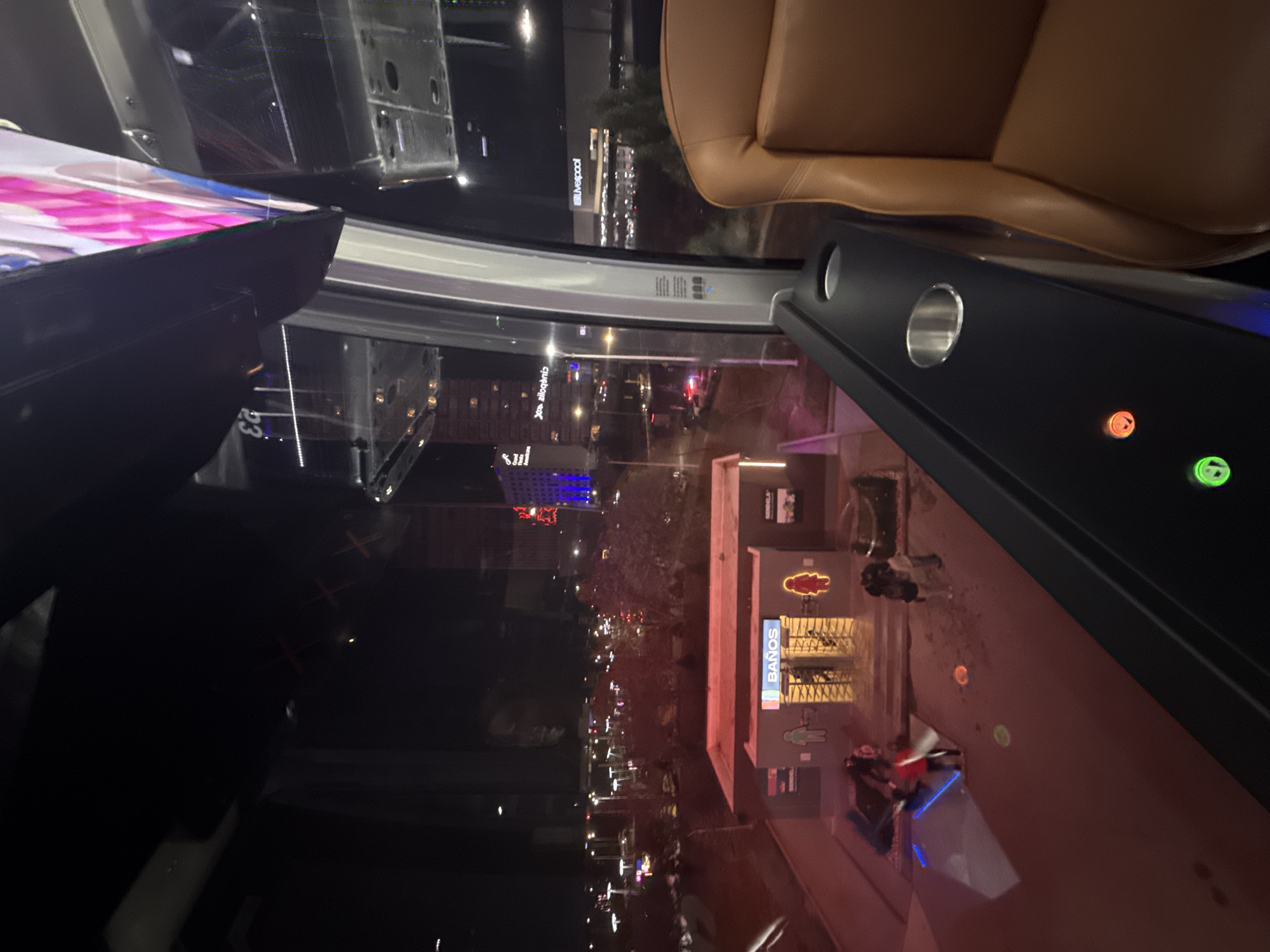 The image, taken from inside a car, shows a nighttime view of the city of Puebla, Mexico. Through the car window, city lights, tall buildings, and street activity are visible. On the right side of the frame, a structure with the word "Baños" illuminated is evident. Outside, there is what appears to be a bathroom. An advertisement for restrooms/toilets is also apparent. In the background, taller buildings and general urban ambiance can be seen. The car's interior is also in the frame, showing a leather seat and a control panel.