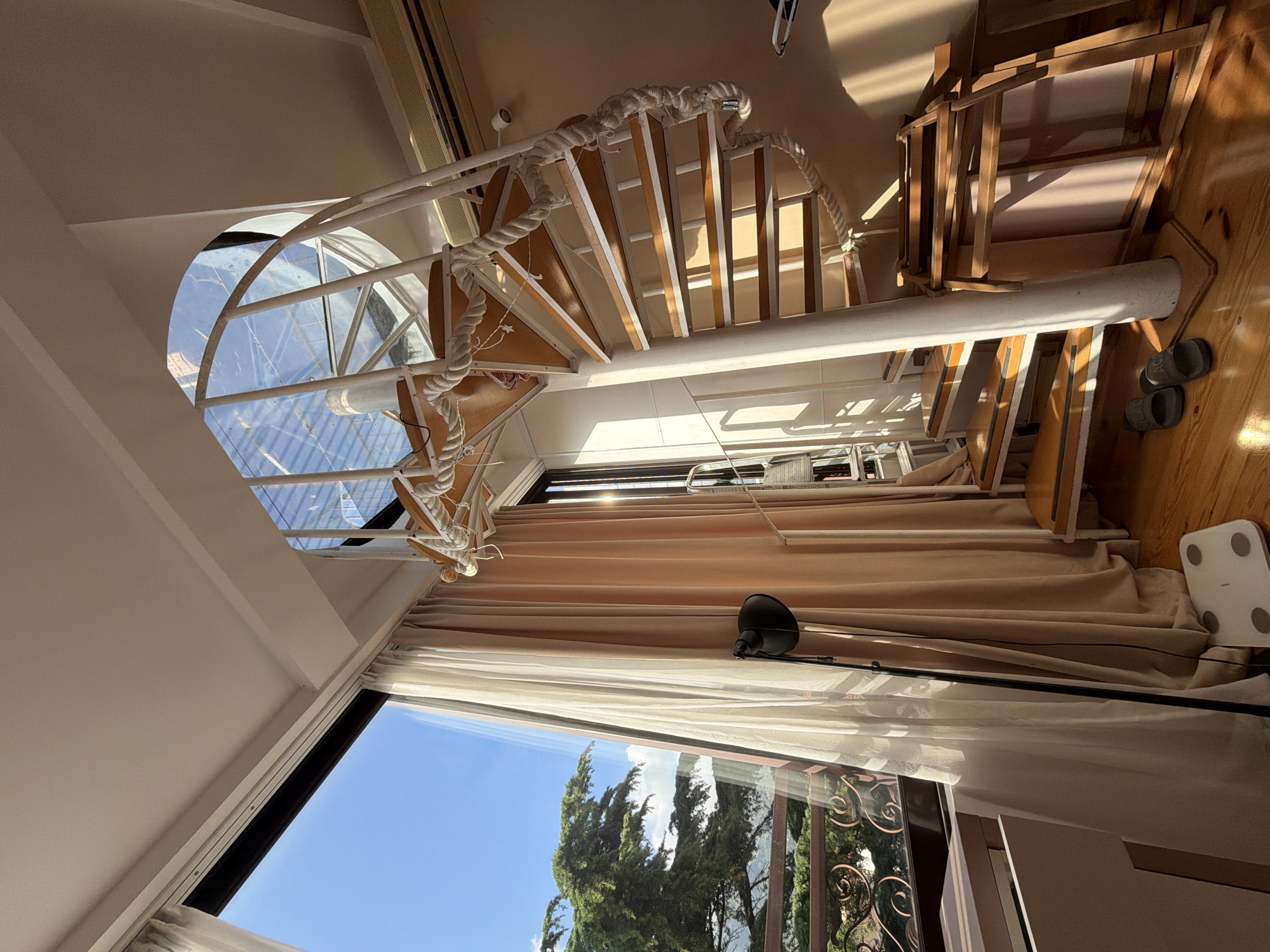 The image shows the interior of a room, likely in a multi-story building. A spiral staircase with a white metal frame and rope detailing leads upwards, its steps partially visible. Light streams in from a curved glass skylight above the stairs. A large window spans the lower portion of the image, revealing a view of tall green trees against a clear blue sky, suggesting an outdoor environment. Beige curtains are draped across the window, partially obscuring the view. On the floor near the window, a black floor lamp is present. To the right, on a wooden floor, there's a bathroom scale and a pair of slippers. The room seems to be well-lit with natural light. The location of the image is Budva, Montenegro. There are no people visible in the image.
