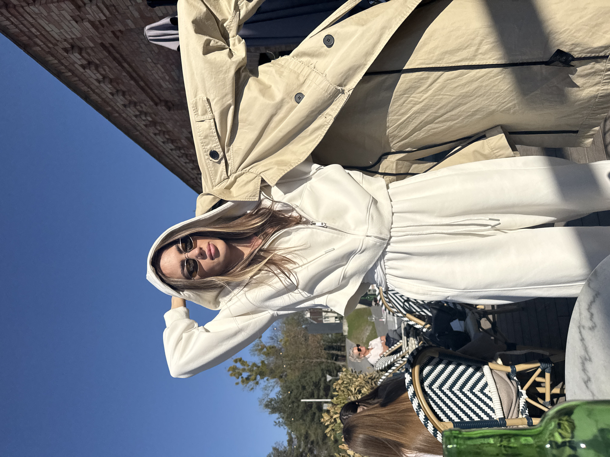 An outdoor, sunlit scene in Warsaw, Poland, features a young woman posing directly for the camera. She is wearing a white hooded tracksuit with a zipper closure and stylish sunglasses, with her light brown hair flowing around her face. Her left hand is raised towards her hood.
Above her, a light-colored, possibly beige or tan, trench coat or jacket is draped or hanging, partially obscuring the background. To her right, a bright blue sky is visible, suggesting clear weather. To her left, a brick building wall is partially seen in the upper background.
The setting appears to be an outdoor cafe or restaurant. Below and to the right of the main subject, a black and white chevron-patterned cafe chair is visible, along with parts of a marble-patterned table and a green glass bottle in the immediate foreground. Another person wearing sunglasses is seated in the mid-ground, and the top of another person's head is visible in the lower right. Trees and general outdoor greenery are present in the distant background. The time of day is bright and sunny, indicating daytime. No visible text is present in the image.