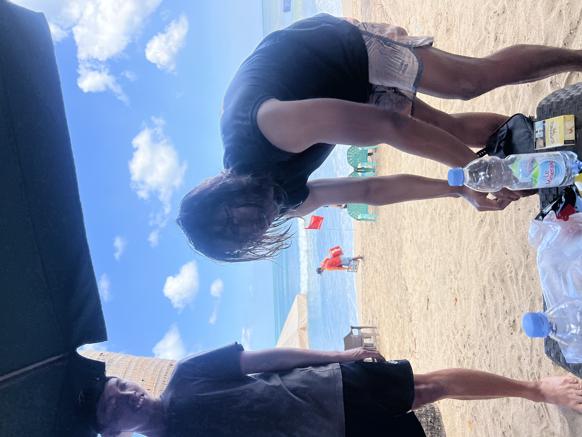 Two young men are at a beach in Kuta, Indonesia. The man on the left, wearing a dark t-shirt and a baseball cap, is smiling. The man on the right, with longer hair and wearing a dark t-shirt and patterned shorts, is bending over, looking down. In front of him, on a woven table, are a bottle of "Le Minerale" water, a pack of "Camel" cigarettes, and other items in a plastic bag. In the background, the ocean waves crash on the sandy shore. A person in an orange shirt is walking towards the water, carrying a red bag. Several green plastic chairs are scattered on the beach, and a beige umbrella shades a portion of the sand. The sky is blue with scattered white clouds.