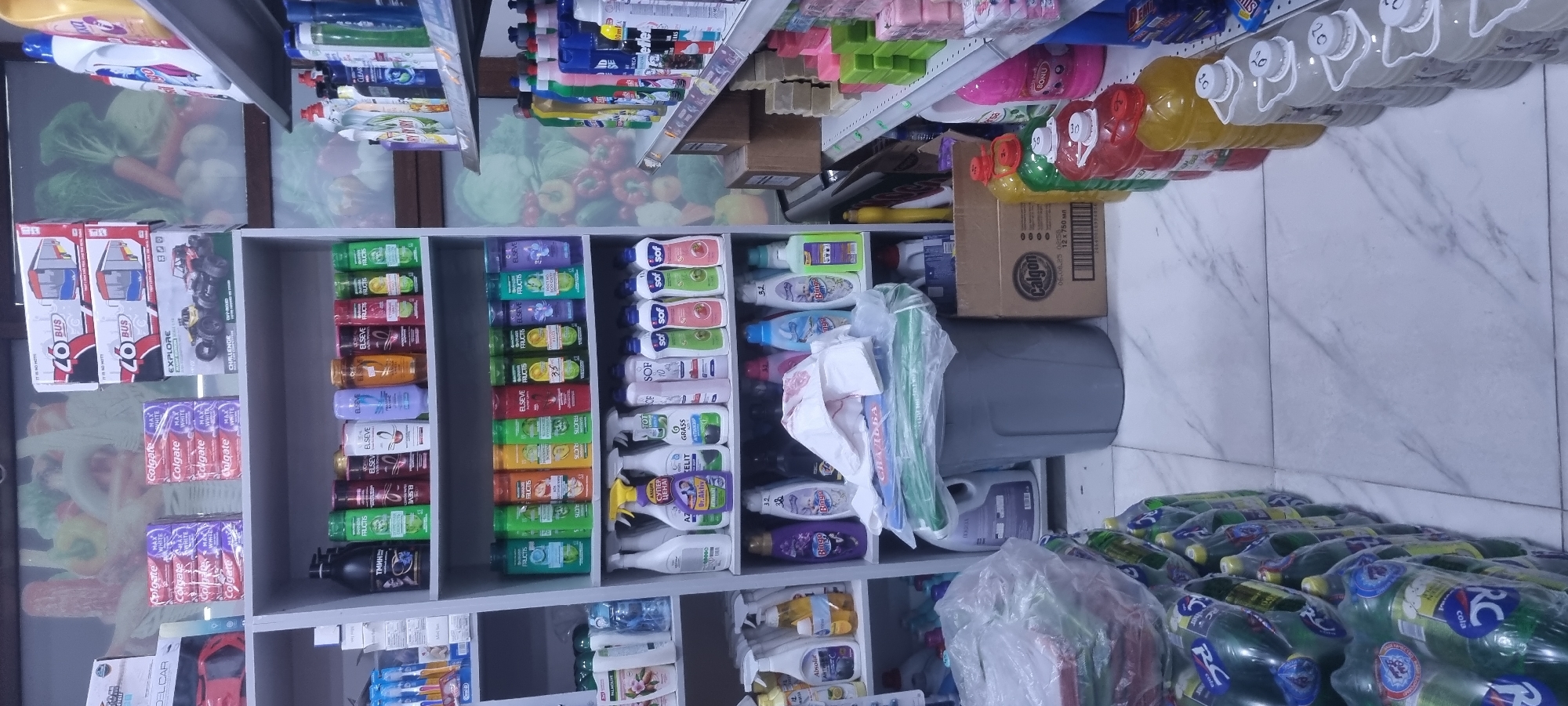 The image shows a retail store interior, likely a section of a supermarket or convenience store, stocked with various household cleaning and personal care products. The shelves are organized and filled with an assortment of bottled liquids, packaged soaps, and boxed items. Prominent brands include Colgate toothpaste, Elseve hair products, and other cleaning agents. In the foreground, stacks of bottled beverages, possibly soft drinks like RC Cola, are visible on the floor. The store appears to be well-lit, with a clean, tiled floor. The overall impression is of a typical retail environment. This scene is located in Ismoili Somoni, Tajikistan.