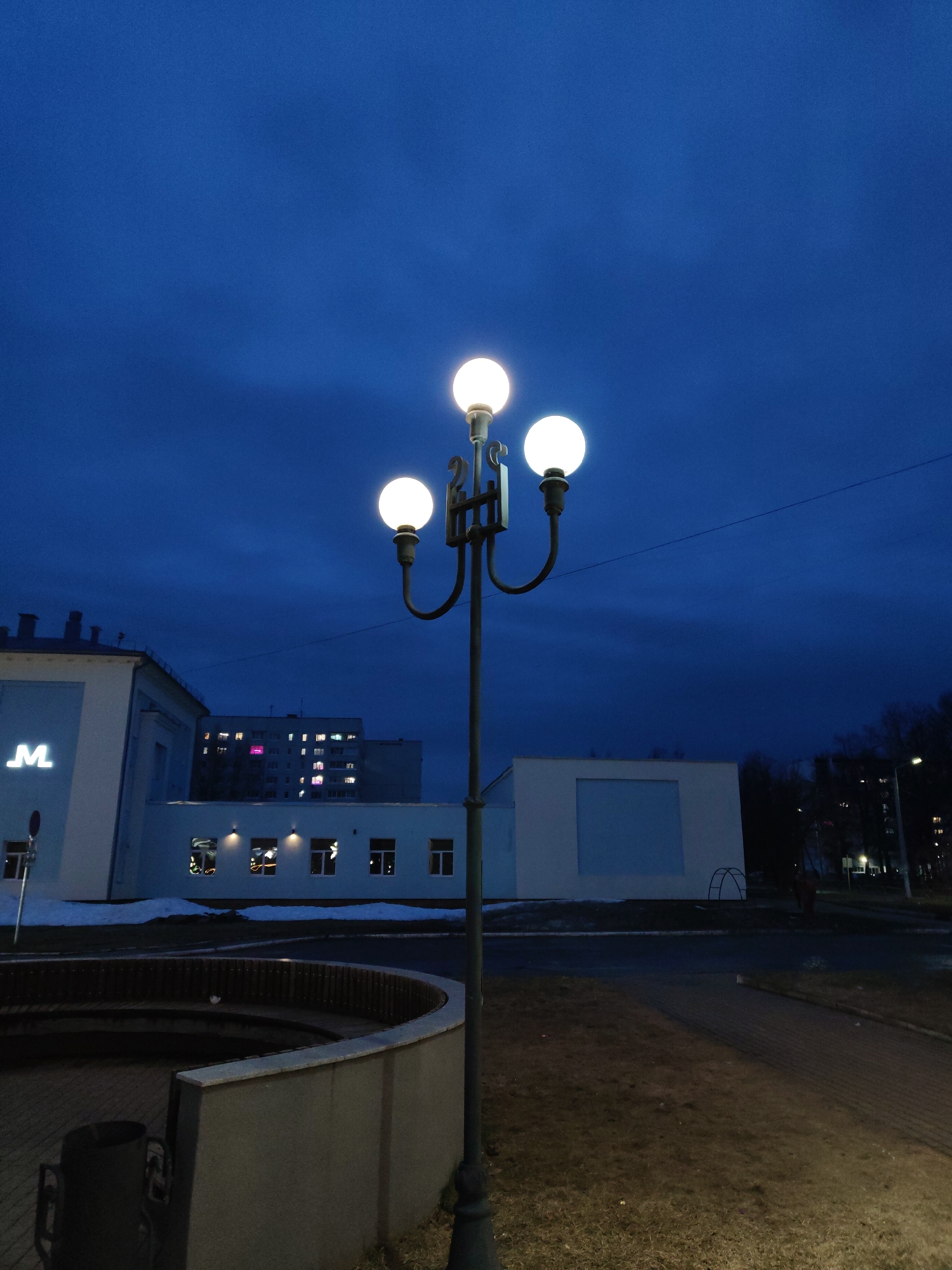 The image shows a street scene at dusk in Unknown, Unknown. A three-bulb street lamp stands prominently in the foreground, illuminated. Behind it is a low, curved concrete structure, possibly a retaining wall or a bench enclosure. In the midground are buildings, including one with a visible "M" logo and a building with several lit windows. Patches of snow are visible on the ground. In the background, a taller building with illuminated windows is visible, along with trees. The sky is a deep blue. The lighting suggests it's either late evening or very early morning. The scene appears quiet with no people visible in clear detail.