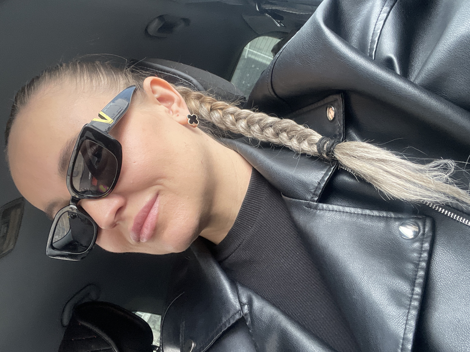 A woman wearing black sunglasses, a black leather jacket, and a black top sits inside a car. Her blonde hair is braided and pulled to one side. The black clover earring is visible on the left ear. The location is most likely not the city of Chaikovsky, Russia as there is no indicator of that in the image.