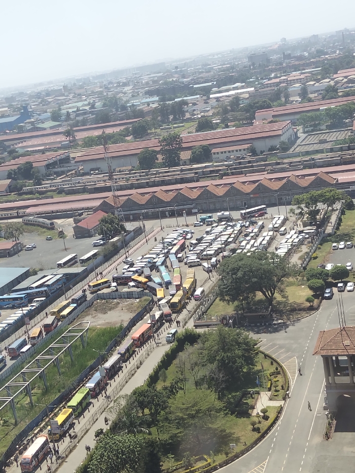 An elevated daytime photograph captures a large urban transport hub adjacent to the Nairobi Railway Station complex in Nairobi, Kenya. The foreground and midground are dominated by a densely packed public transport terminal, containing hundreds of vehicles. These vehicles are primarily brightly colored matatus (minibuses) and a lesser number of larger buses, arranged in tight rows across multiple asphalt parking bays and queuing lanes. Numerous pedestrians are observed walking between the vehicles and along paved areas. A long queue of matatus extends along a road section to the left, bordered by a fence. To the right, a manicured green space with trees and a roundabout is visible, adjacent to a brown-roofed structure resembling a guardhouse. The background features the extensive infrastructure of the Nairobi Railway Station, including multiple parallel railway tracks with numerous parked train carriages (both passenger and freight), and several large industrial-style buildings with distinctive reddish-brown pitched roofs. A tall metal lattice tower stands within the railway complex. The distant background shows a dense urban sprawl under a hazy sky.