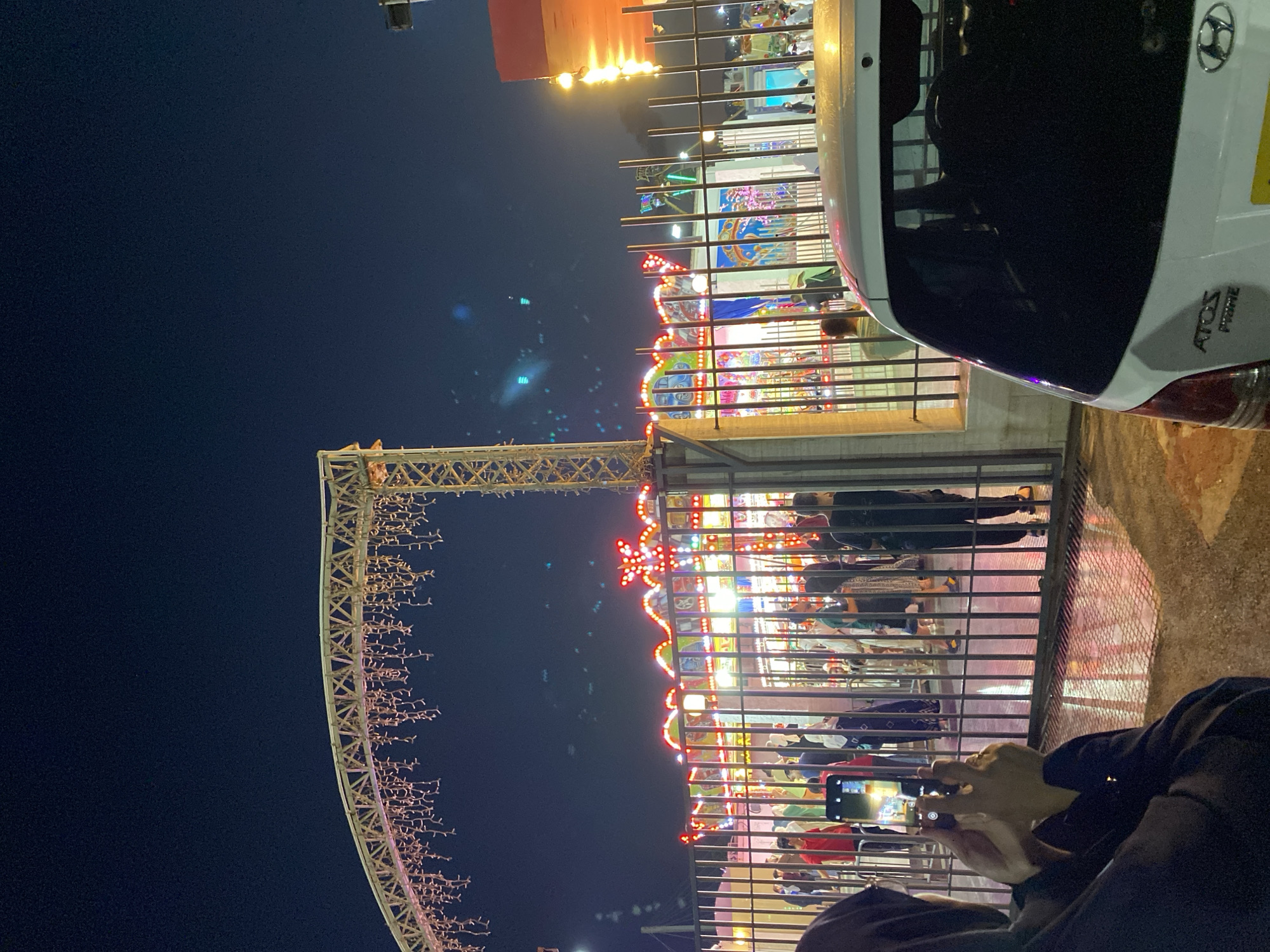 In the city of Hussein Dey, Algeria, at night, a group of people stand behind a black metal fence, watching a brightly lit amusement ride. The ride is decorated with colorful lights and appears to be a carousel. A person in the foreground on the left is holding up a smartphone, taking a picture of the scene. To the right, the rear of a white Hyundai Atos Prime car is visible. The overall atmosphere suggests a festive event, possibly a fair or carnival, taking place after dark.