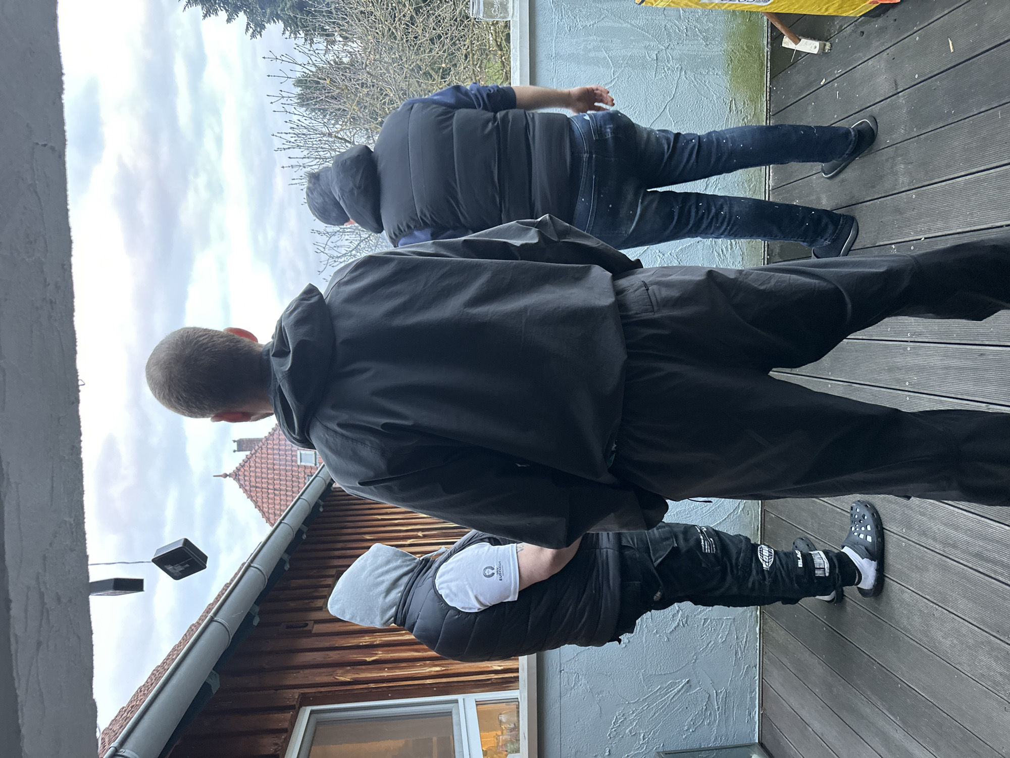 Three people are standing on a wooden deck. The first person, closest to the camera, is facing away from the camera and wearing black pants, black shoes, and a black hooded jacket. The second person is facing away from the camera and wearing jeans, black shoes, and a black hooded vest. The third person is wearing black pants, black shoes, a black vest, and a gray hood. They are standing next to a house with a brown roof. The location is Heinebach, Germany.