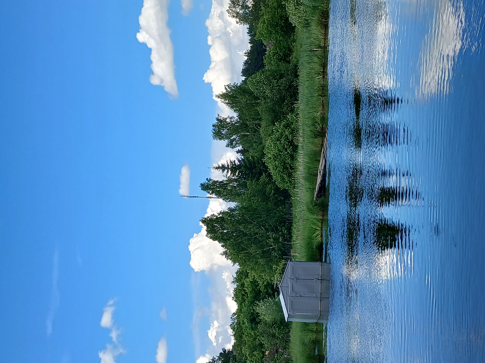 The image depicts a serene waterside scene with a lush green shoreline under a bright blue sky dotted with white clouds. On the water, near the edge of the bank, sits a small, gray shed with a silver roof. The water's surface is rippled, reflecting the sky, clouds, and the dense green foliage that lines the bank. A wooden structure, possibly a dock or a pier, is partially visible extending into the water on the right side of the frame. The overall impression is one of a tranquil, natural setting, likely during the daytime given the brightness of the sky and the clear reflections. The location context suggests this scene is in Yekaterinburg, Russia. There is no visible text in the image.