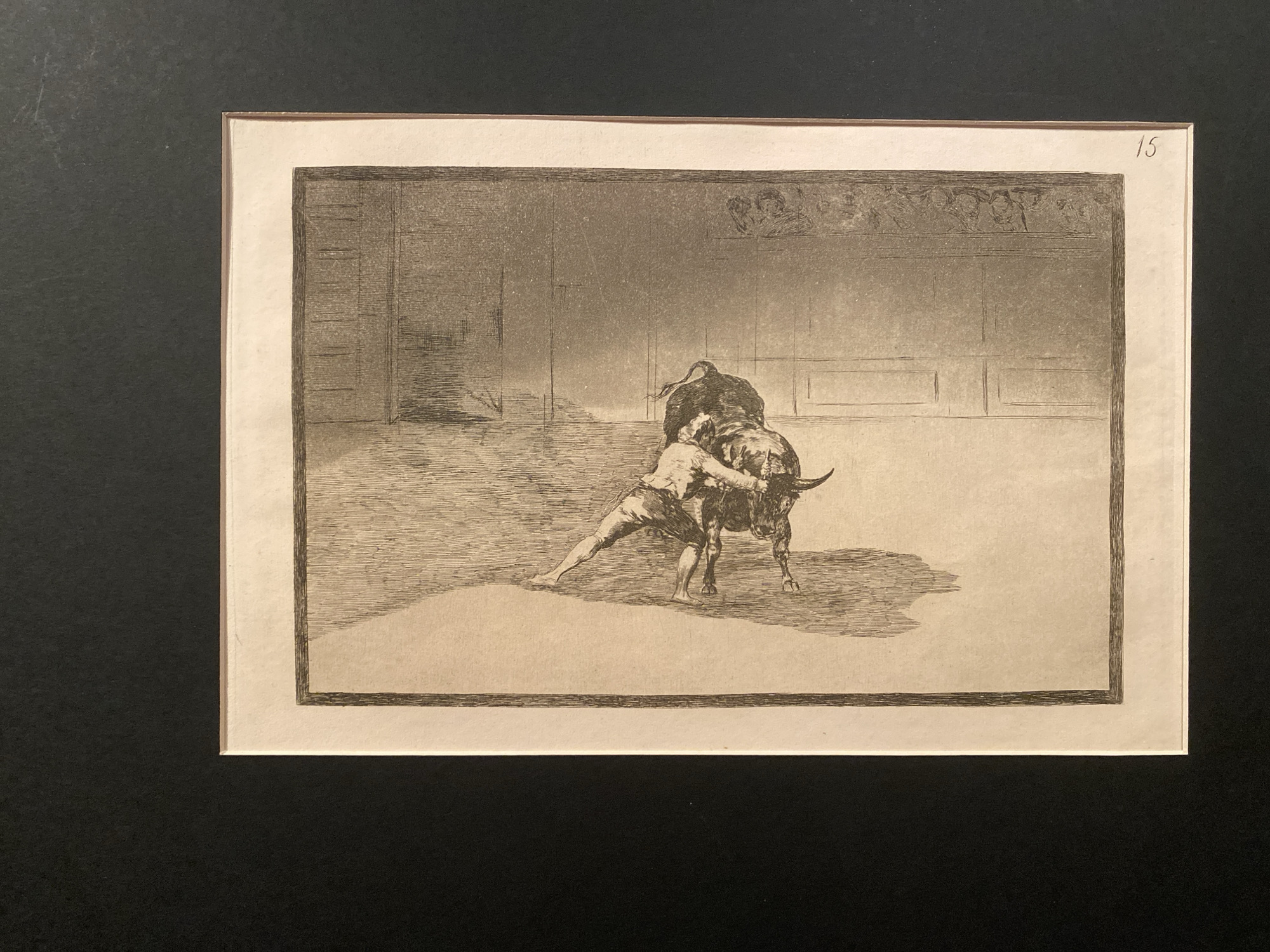 The image is a framed etching depicting a bullfight. In the center, a man is grappling with a bull, leaning into its neck and head. The man is dressed in simple trousers and a shirt, his body strained with effort. The bull is muscular and appears to be pushing against the man. The scene takes place in an arena with a dirt floor and a wall in the background that has some decorative elements at the top and vertical panels lower down. The etching is matted and framed, with the number "15" handwritten in the upper right corner of the mat. The artwork is presented against a dark background, likely a wall or display surface. There is no indication of the city of Sofia, Bulgaria, or any specific location within it.