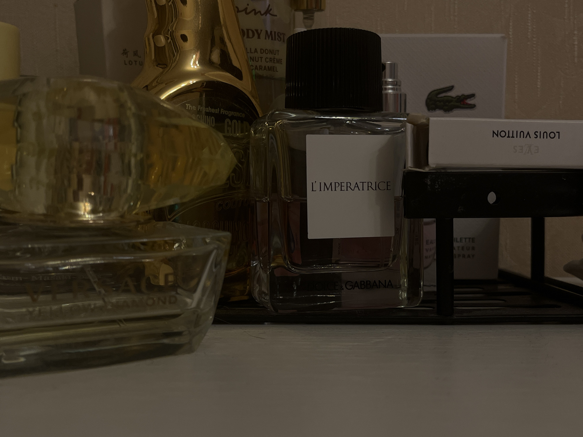 An indoor, close-up, eye-level, medium shot captures a collection of various perfumes displayed on a white surface. The lighting is dim, casting shadows and highlighting the reflective surfaces of the perfume bottles. In the foreground, a clear glass perfume bottle with a faceted design and a yellow liquid is positioned on the left. To its right, a taller, clear glass perfume bottle with a black cap and a white label that reads "L'IMPERATRICE" is prominent. Behind this bottle, partially obscured, is a large, gold-colored perfume bottle. Further in the background, a white box with a green alligator logo, a white box with the words "LOUIS VUITTON" and "EYES" are visible. A black metal rack supports some of the bottles. The overall environment suggests a vanity or display shelf.