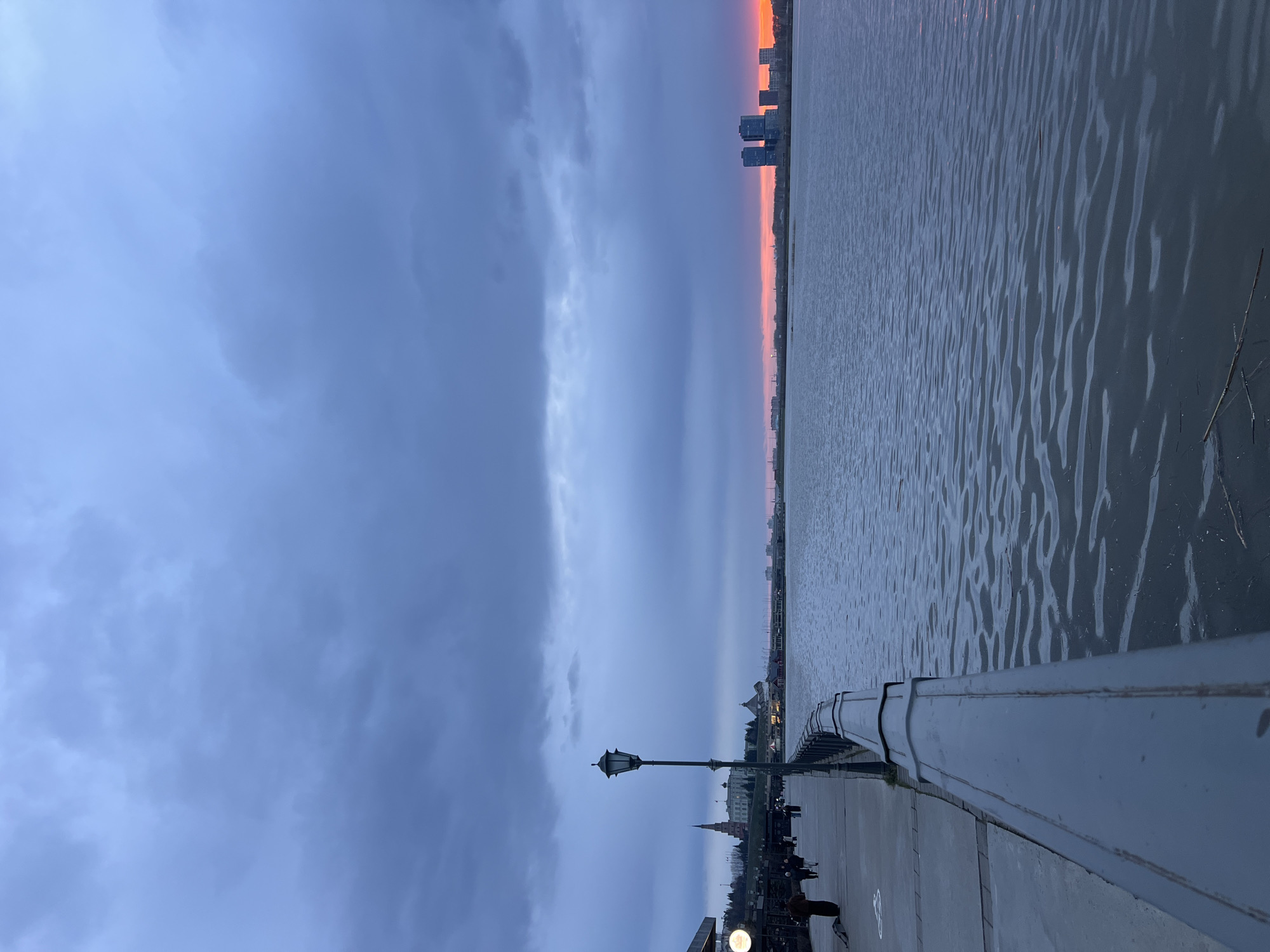 The image shows a dusk scene on the Kazan Kremlin Embankment, overlooking the Kazanka River in Kazan, Russia. The sky is overcast with dark clouds, though an orange glow is visible on the horizon, possibly from the setting sun. The Kazanka River stretches out towards the horizon with buildings visible in the distance. A grey railing runs along the embankment's edge. There is a lamppost with a vintage design. A group of people can be seen walking and standing along the embankment.