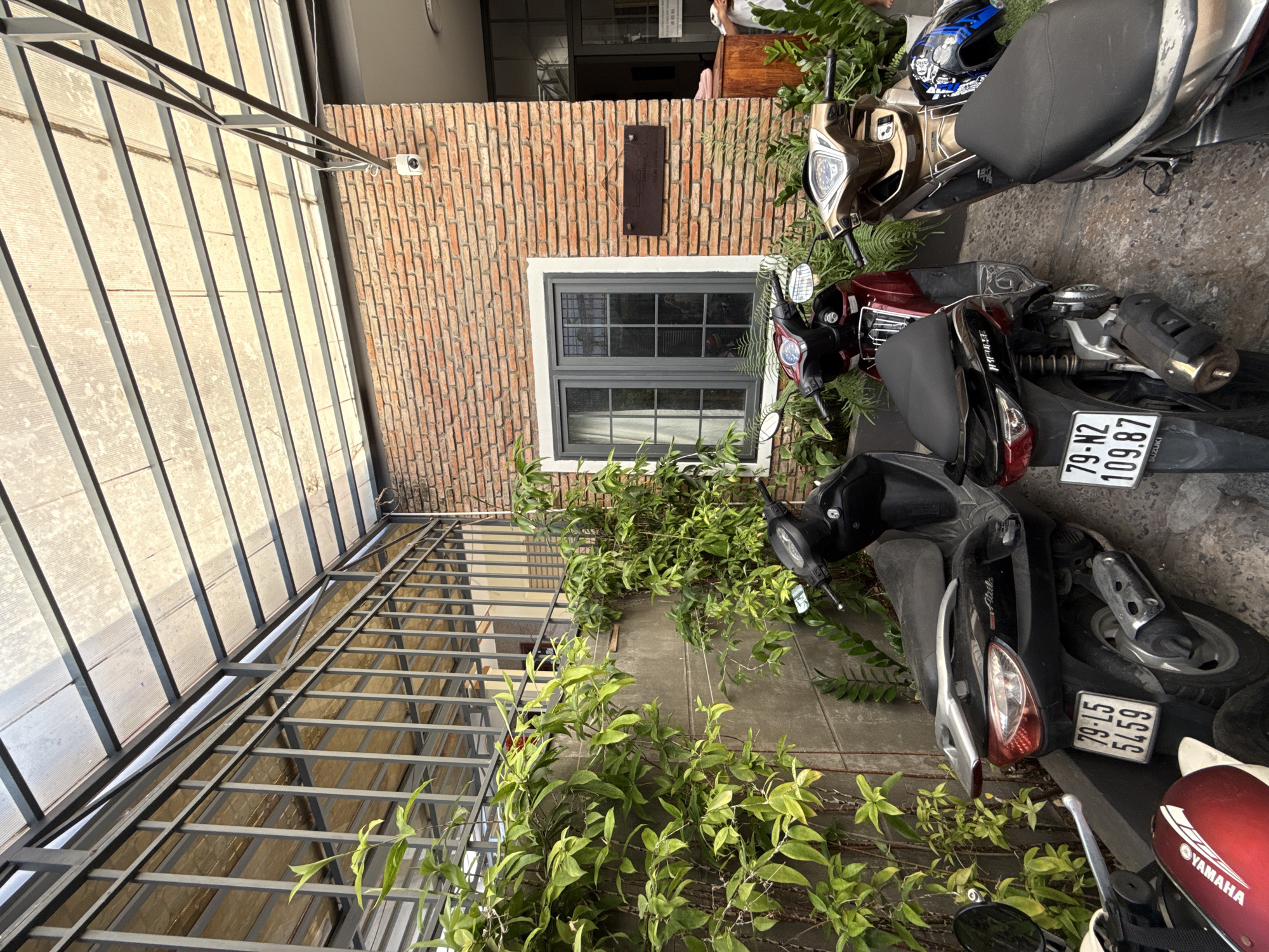 The image depicts a narrow alleyway or passage in Nha Trang, Vietnam, with several parked motorcycles. The scene is partially covered by a corrugated polycarbonate roof supported by a metal frame. To the left, a textured concrete wall is covered with lush green foliage, including vines and ferns. A metal security fence with vertical bars runs along the top of the wall, obscuring the view of what lies beyond.
In the foreground, three motorcycles are parked side-by-side. The leftmost is a red Yamaha. Next to it is a black scooter with a license plate reading "79-L5 5459". To the right is a bronze scooter with a license plate reading "79-N2 109.87". A blue and black helmet rests on the seat of the bronze scooter.
The background features a building with a brick facade. A prominent feature of the building is a rectangular window with a dark gray frame and multiple panes. Above the window, a security camera is visible mounted on the brick wall. To the right of the window, a dark wooden sign with white lettering hangs on the brick wall. Further to the right, a doorway leads into the building, with a glimpse of a reception area or interior space. A person is partially visible in the doorway. The ground is paved with grey tiles or concrete. The overall impression is that of a residential or commercial property entrance with limited parking space.