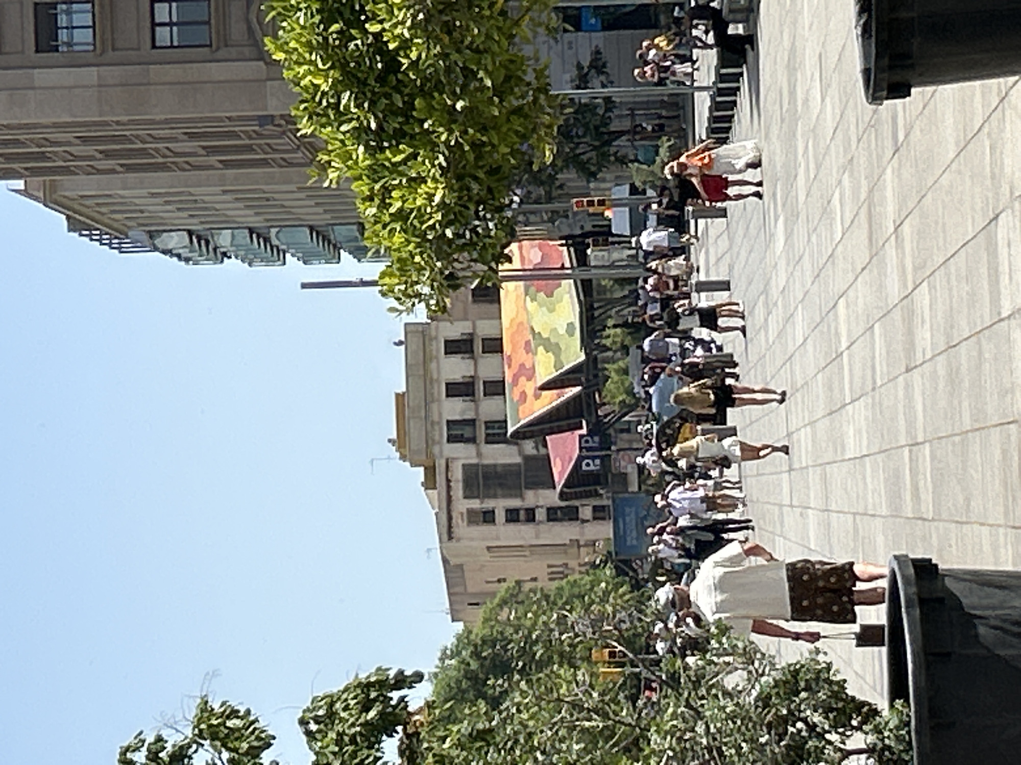 The image depicts a sunny day on a wide pedestrian walkway in Barcelona, Spain, likely near La Rambla or a similar bustling urban area. The walkway is paved with light-colored square tiles and lined with trees on the left. Several people are walking in both directions. On the left, a man wearing a white short-sleeved shirt and patterned shorts walks away from the camera, carrying a small dark bag in his right hand. To his right, a group of people are walking together, some in casual summer clothing.
In the background, buildings of varying heights and architectural styles are visible. The most prominent structure in the mid-ground is a market-like building with a distinctive multi-colored, tent-like roof featuring geometric patterns of orange, yellow, and green. Further back, a tall, modern building with many windows stands to the right, and a more traditional multi-story building with balconies is visible behind trees on the left. The sky is clear blue with no clouds.
The scene suggests a lively tourist area with people enjoying the outdoor space and architecture. The presence of benches and large trash receptacles indicates a public thoroughfare designed for pedestrian traffic. The overall atmosphere is one of urban activity and leisure under bright sunlight.
