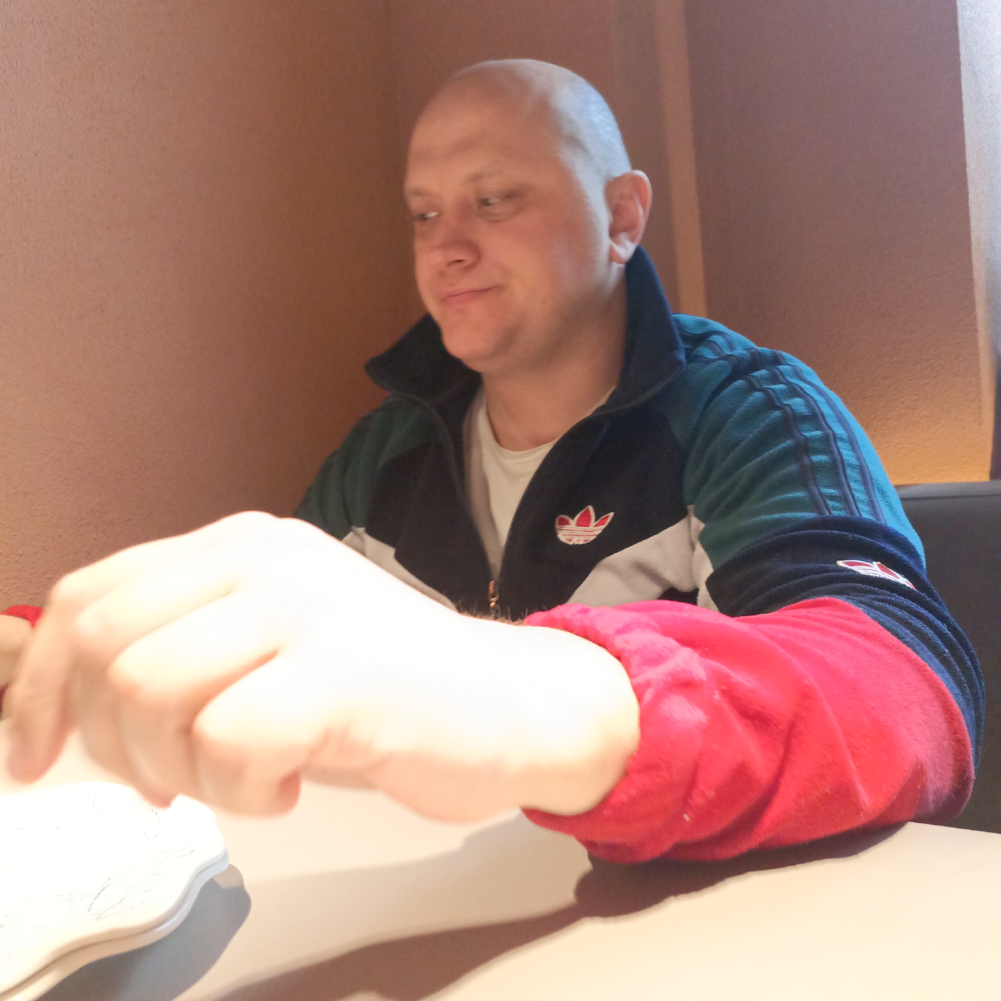 A mid-shot captures a bald man seated at a table in an indoor setting, likely a cafe or casual restaurant, with a textured reddish-brown wall forming the background.
The man, appearing to be in his 30s or 40s, has a light complexion and a clean-shaven head. He is looking downwards and slightly to his right, with a subtle smirk or contemplative expression. His left hand, partly out of focus, is positioned near a white, irregular object, possibly a plate or coaster, on the light-colored table surface.
He is dressed in a multi-colored track jacket over a white t-shirt. The jacket features a dark blue or black collar and upper body, teal or dark green sleeves with visible black stripes, white horizontal sections across the chest, and bright red lower sleeves and cuffs. A distinctive white and red Adidas trefoil logo is embroidered on the left chest of the jacket. A smaller, similar logo is also visible on the cuff of his right sleeve. The jacket is partially unzipped, revealing the white t-shirt underneath.
The lighting indicates an indoor daytime environment, casting soft shadows on the table. The man's posture and expression suggest a calm and relaxed mood.